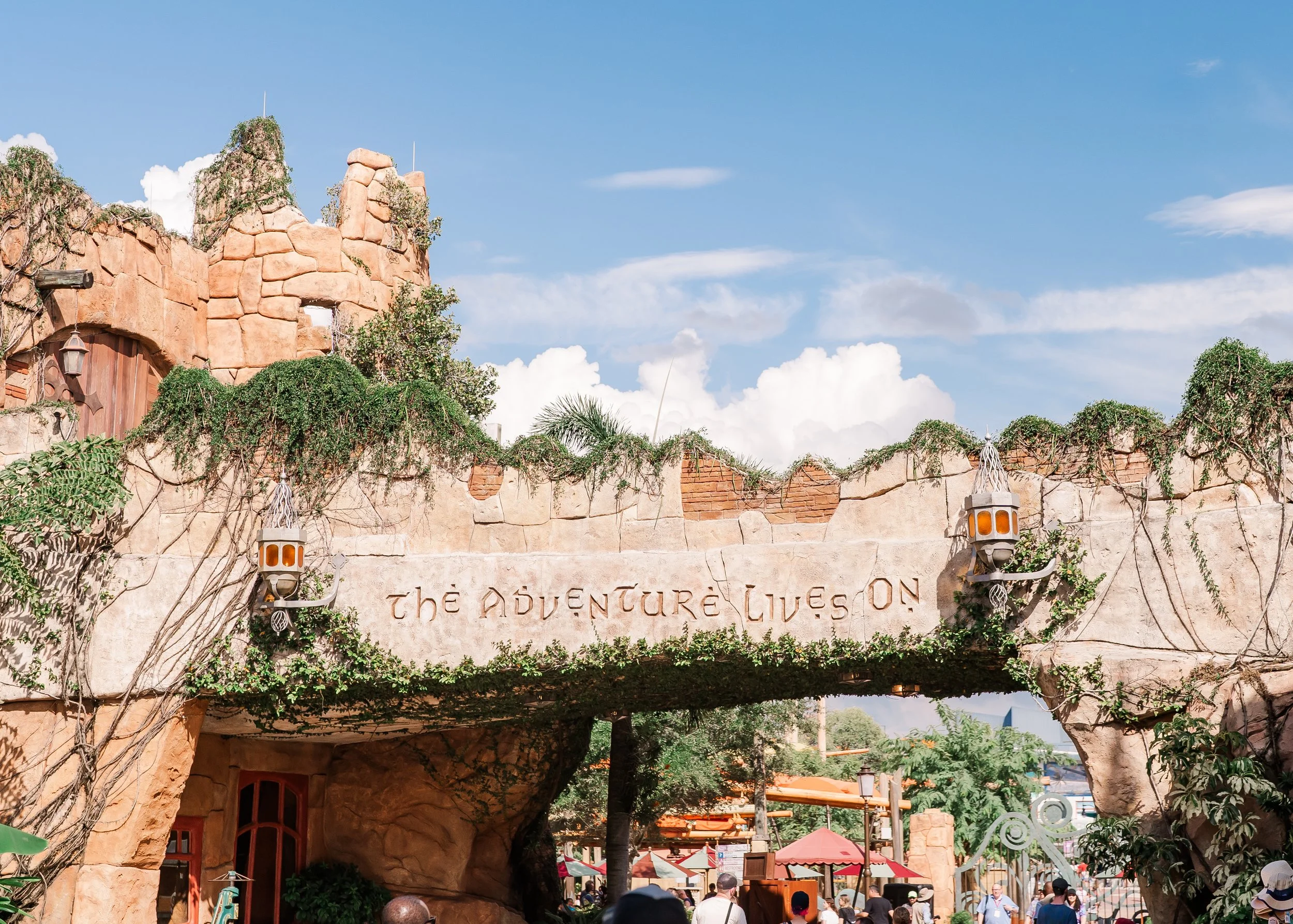 Theme park attraction with rock-like structures, greenery, and a sign stating 'The Adventure Lives On' amid visitors and trees under a blue sky with clouds.