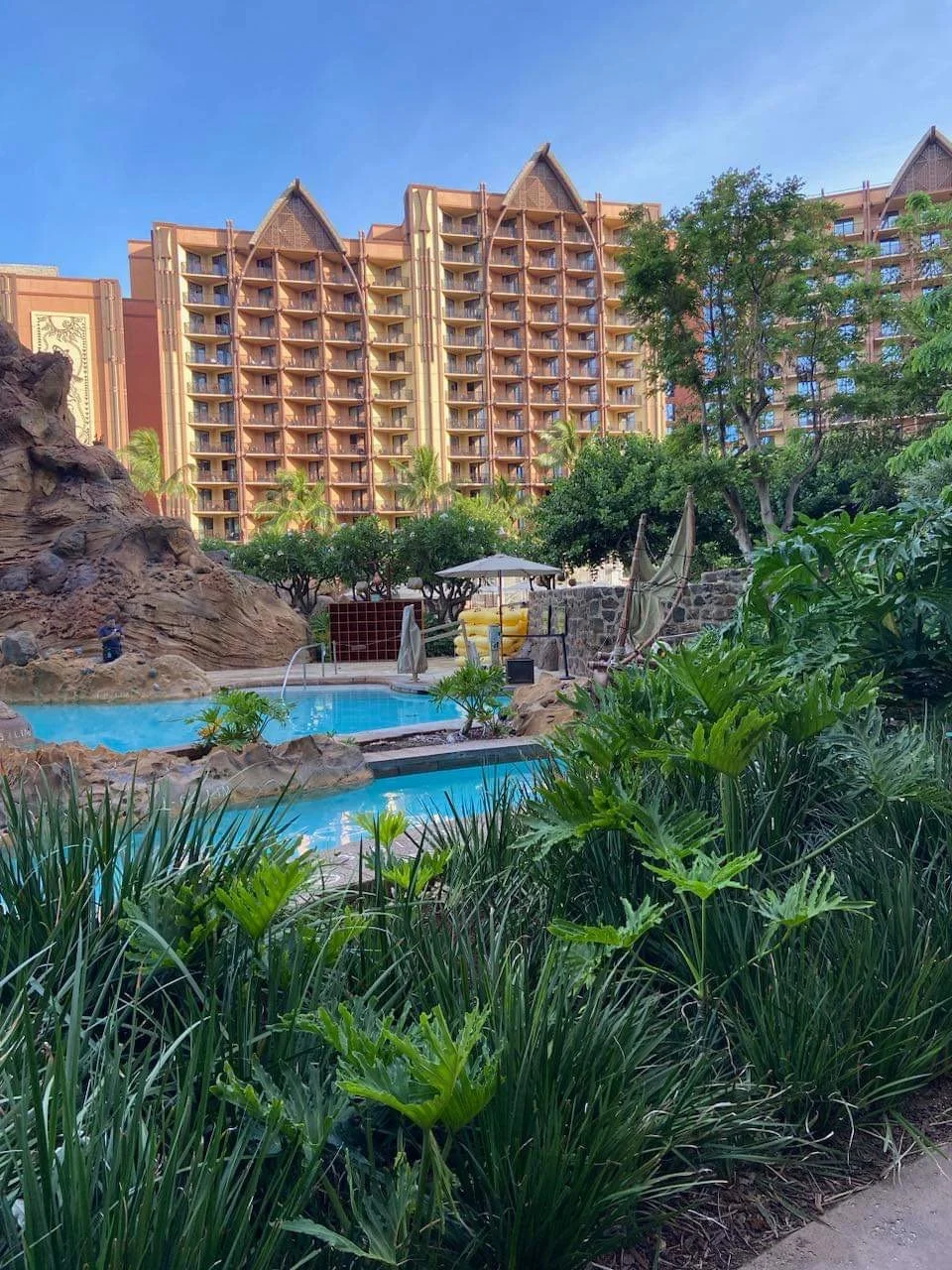 View of a resort with a swimming pool surrounded by lush greenery, lounge chairs, and umbrellas in front of a tall, beige building with multiple balconies and tropical trees.