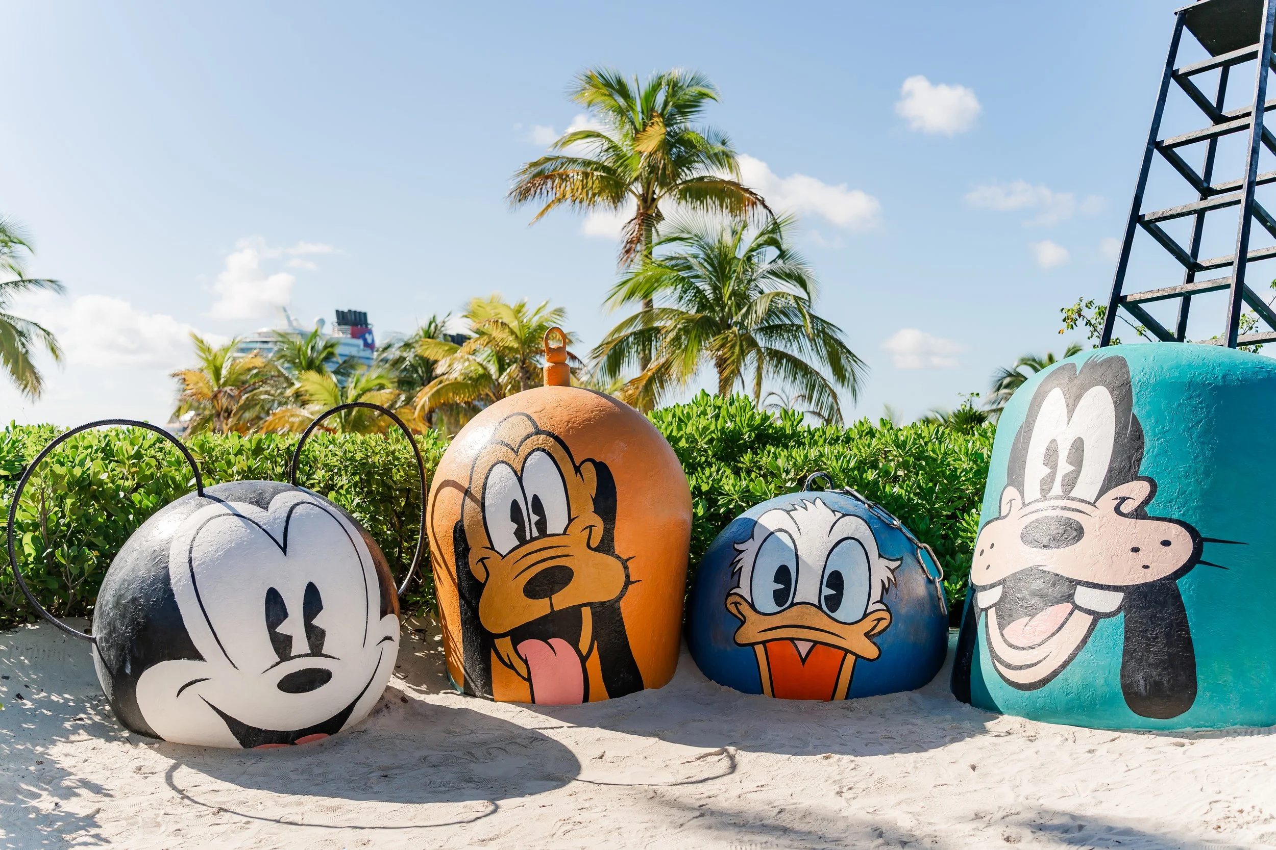 Colorful beach art sculptures of Disney characters Mickey Mouse, Pluto, Daisy Duck, Goofy, and others on sandy shore with palm trees and a cruise ship in the background.