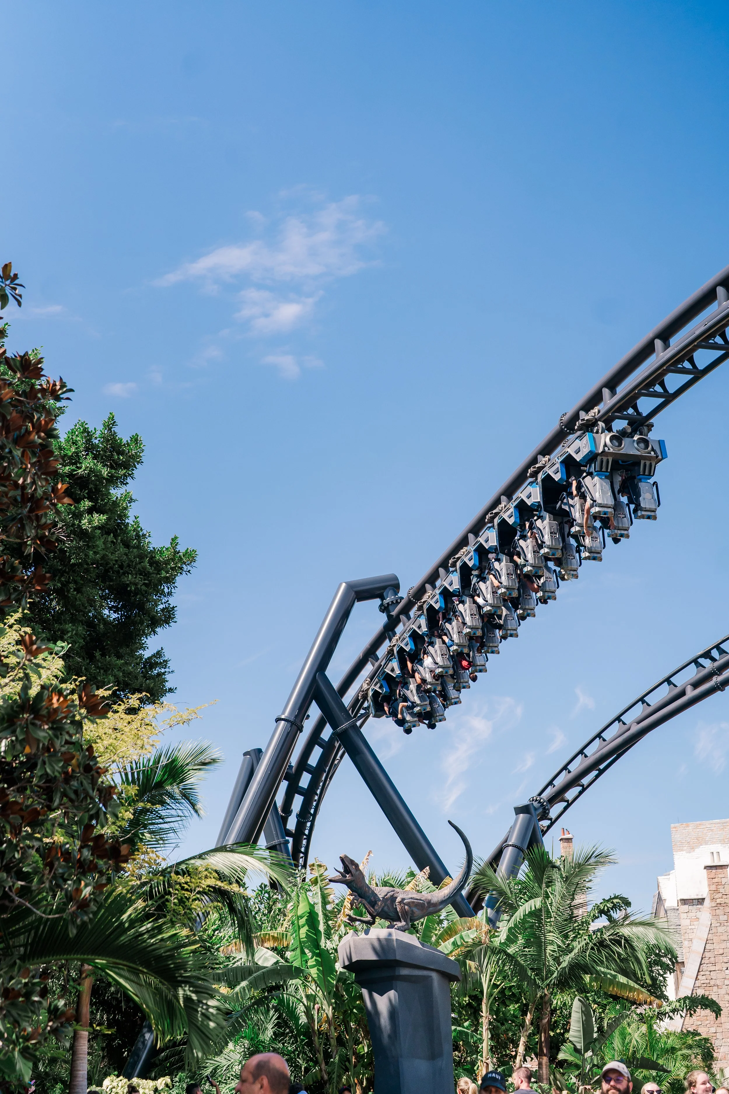 A roller coaster track with multiple lit-up cars passing overhead against a bright blue sky with a few clouds, surrounded by green trees and some people visible at the bottom of the image.