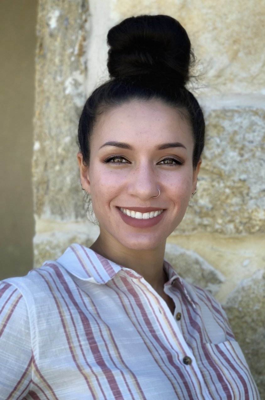 A young woman with dark hair in a high bun, wearing a striped button-up shirt, smiling with a stone wall in the background.