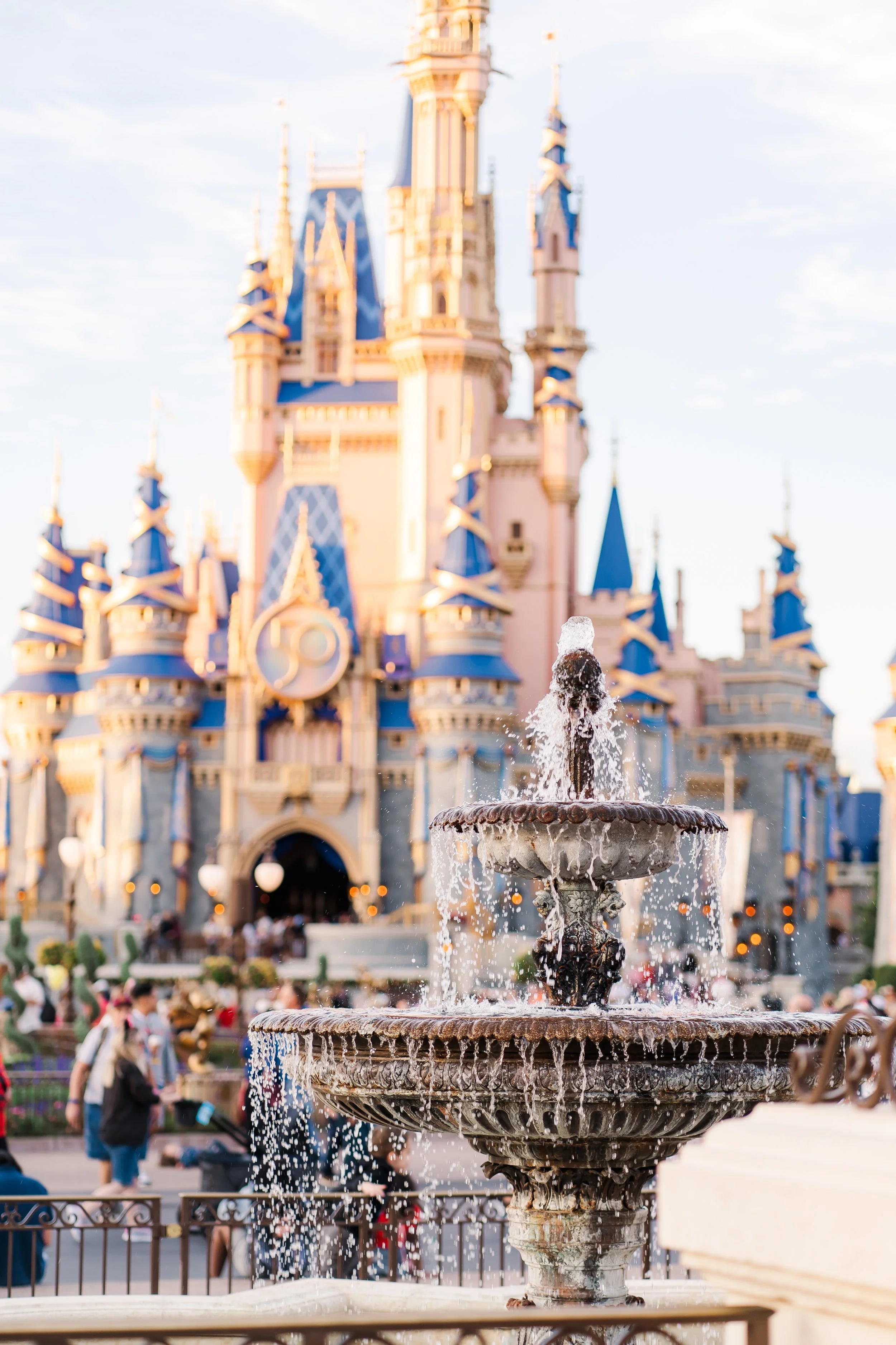 A decorative fountain with water flowing from its top, positioned in front of a castle with blue turrets and pink walls, bustling with visitors in a theme park.