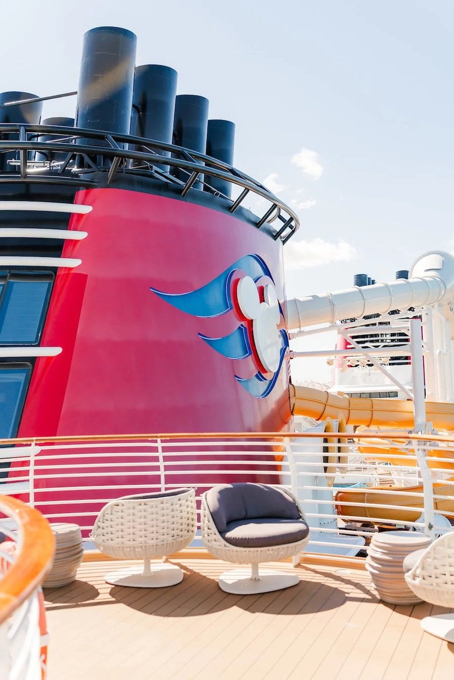 On the deck of a cruise ship, colorful water slides and a large ship's smokestack with a cartoonish face logo.
