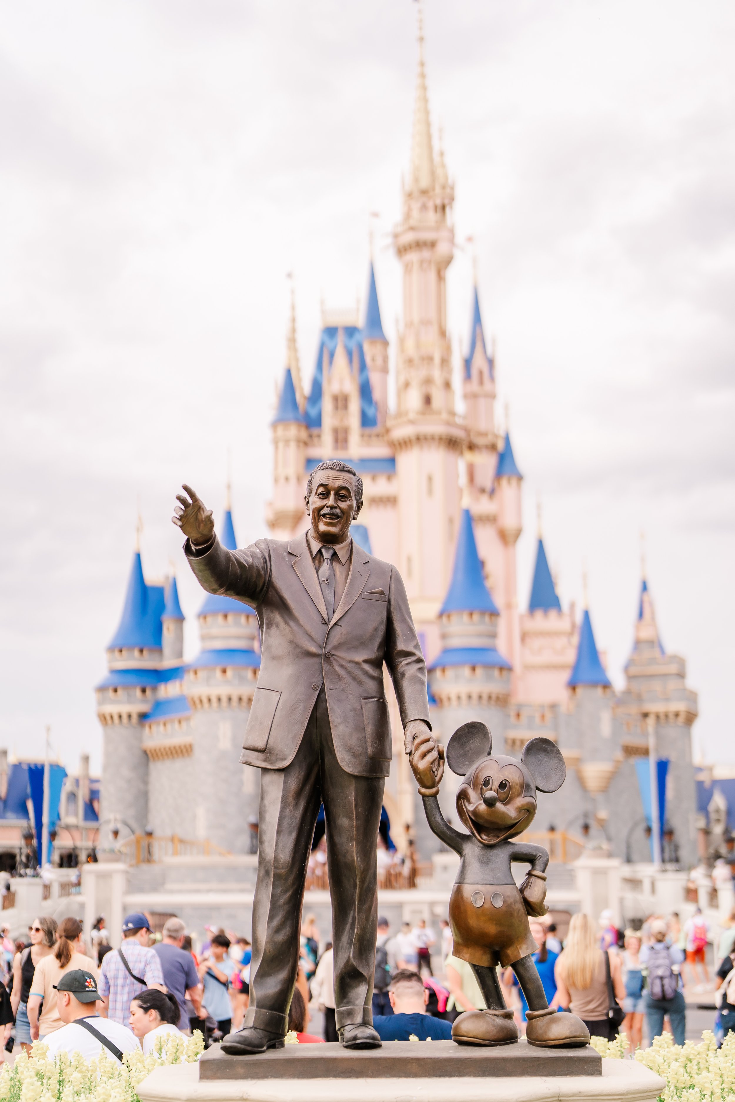 Statue of Walt Disney and Mickey Mouse in front of a castle at Disneyland, with visitors in the background.