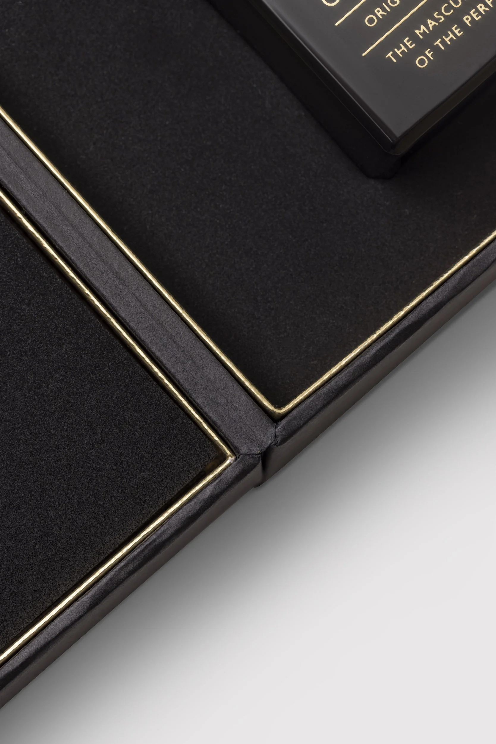 Close-up of a black gift box with a gold-lined border, featuring a black rectangular box inside with gold lettering on top.
