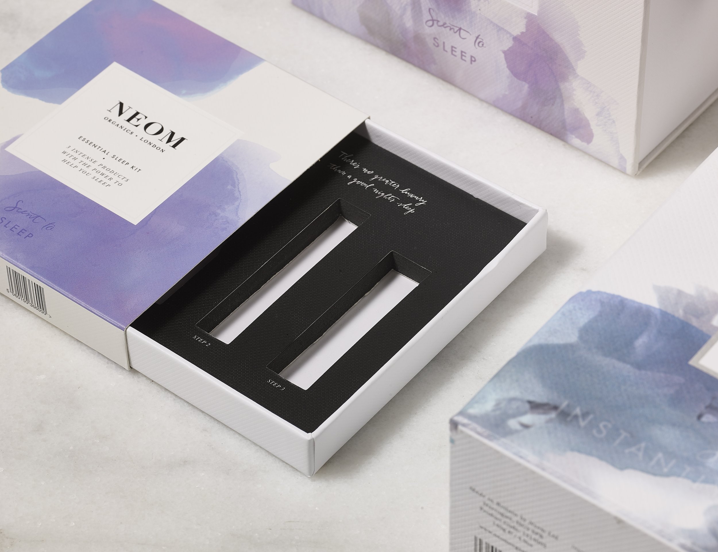 A sleep kit box with a purple and white watercolor design, labeled 'Sent to Sleep,' partially open revealing a black interior section. A promotional card with the text 'NEOM Organics London Essential Sleep Kit - 3 Essential Products with the Power to Help You Sleep' and a decorative watercolor background is visible beside the box on a white surface.