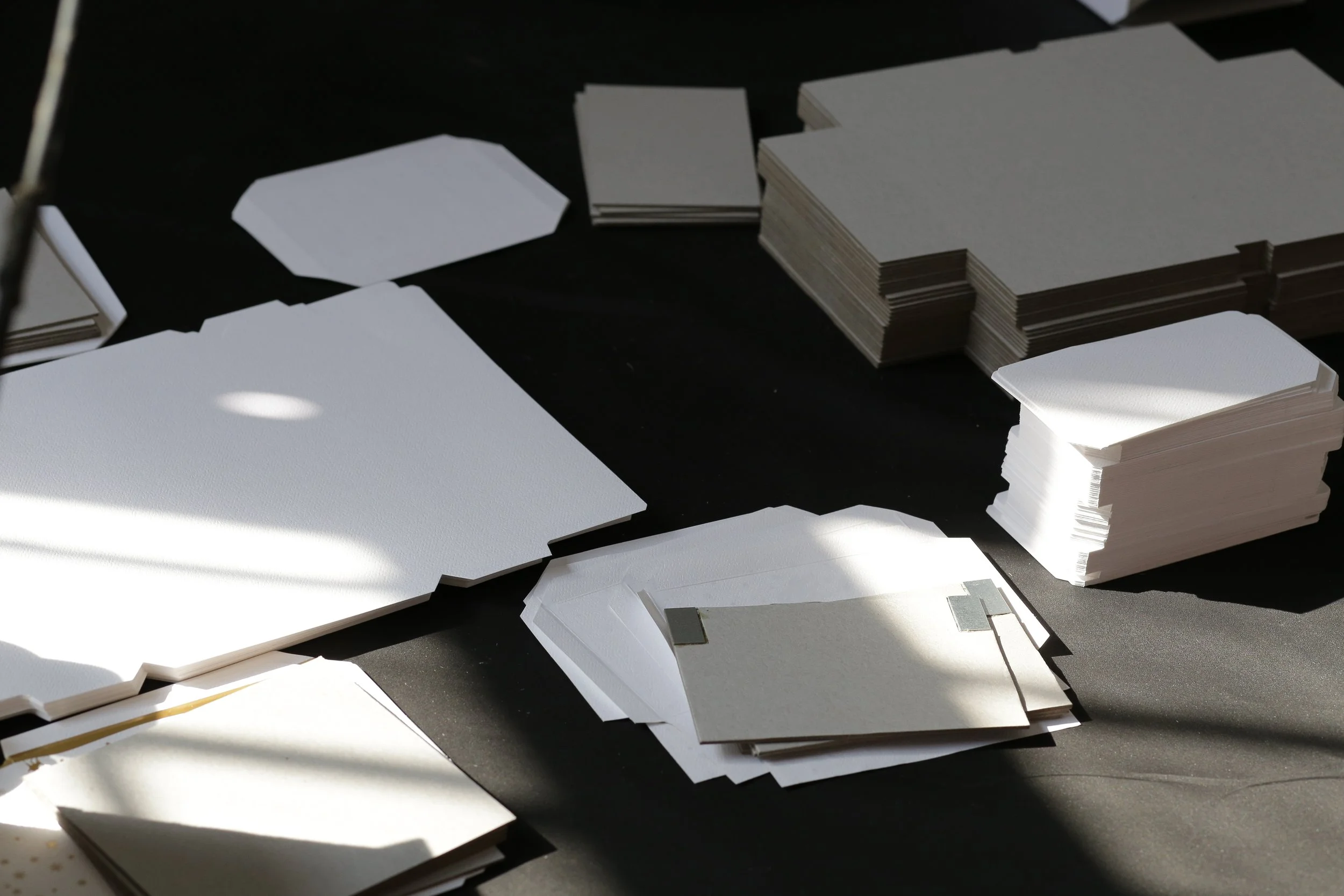 Numerous stacks of blank white and gray papers, some cut into geometric shapes, spread out on a dark surface with sunlight casting shadows.