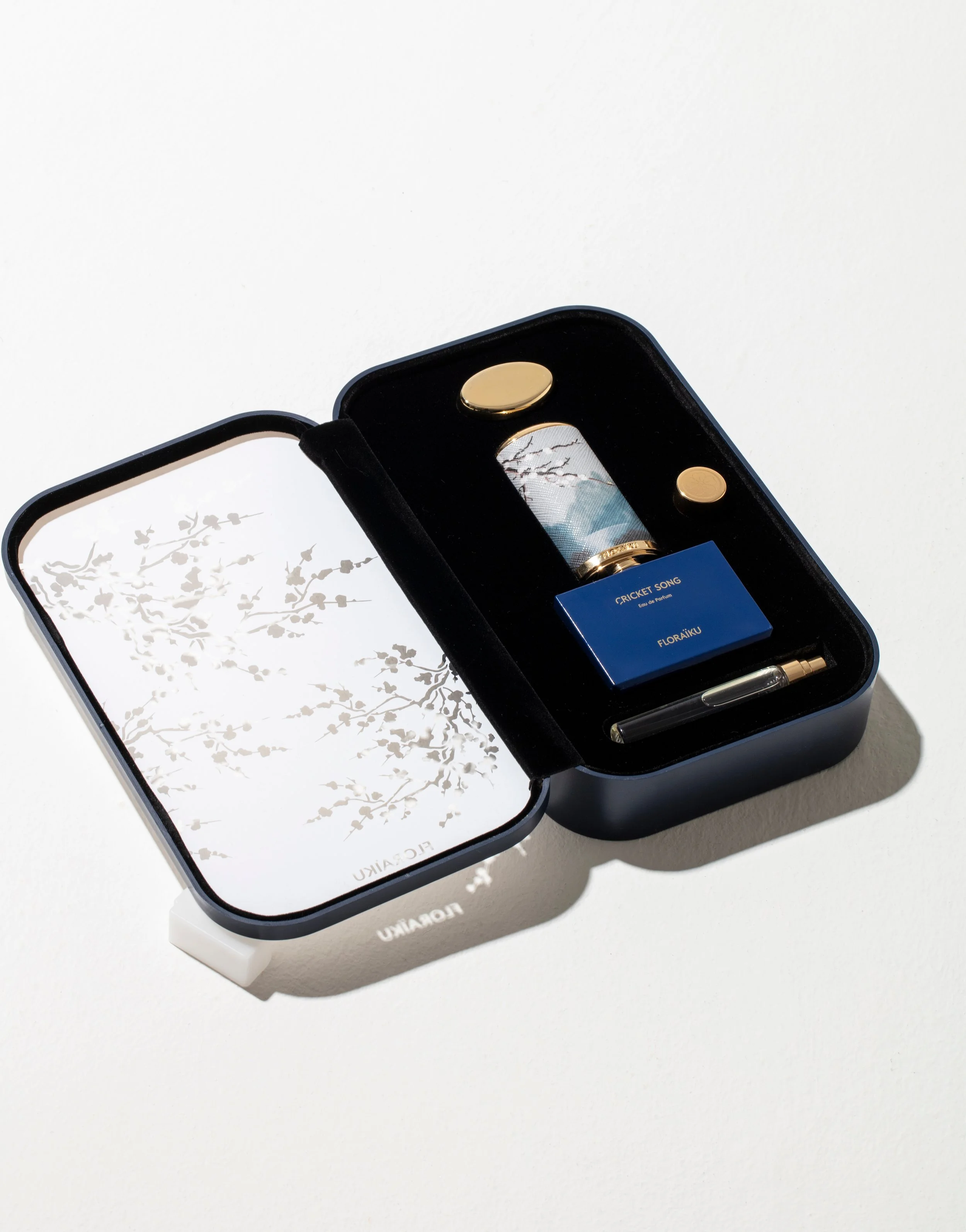 Open black box containing a blue perfume bottle labeled 'Cricket Song' by FlorAiku, a small gold item, and a pen, with white interior decorated with a branch and bird silhouette design.