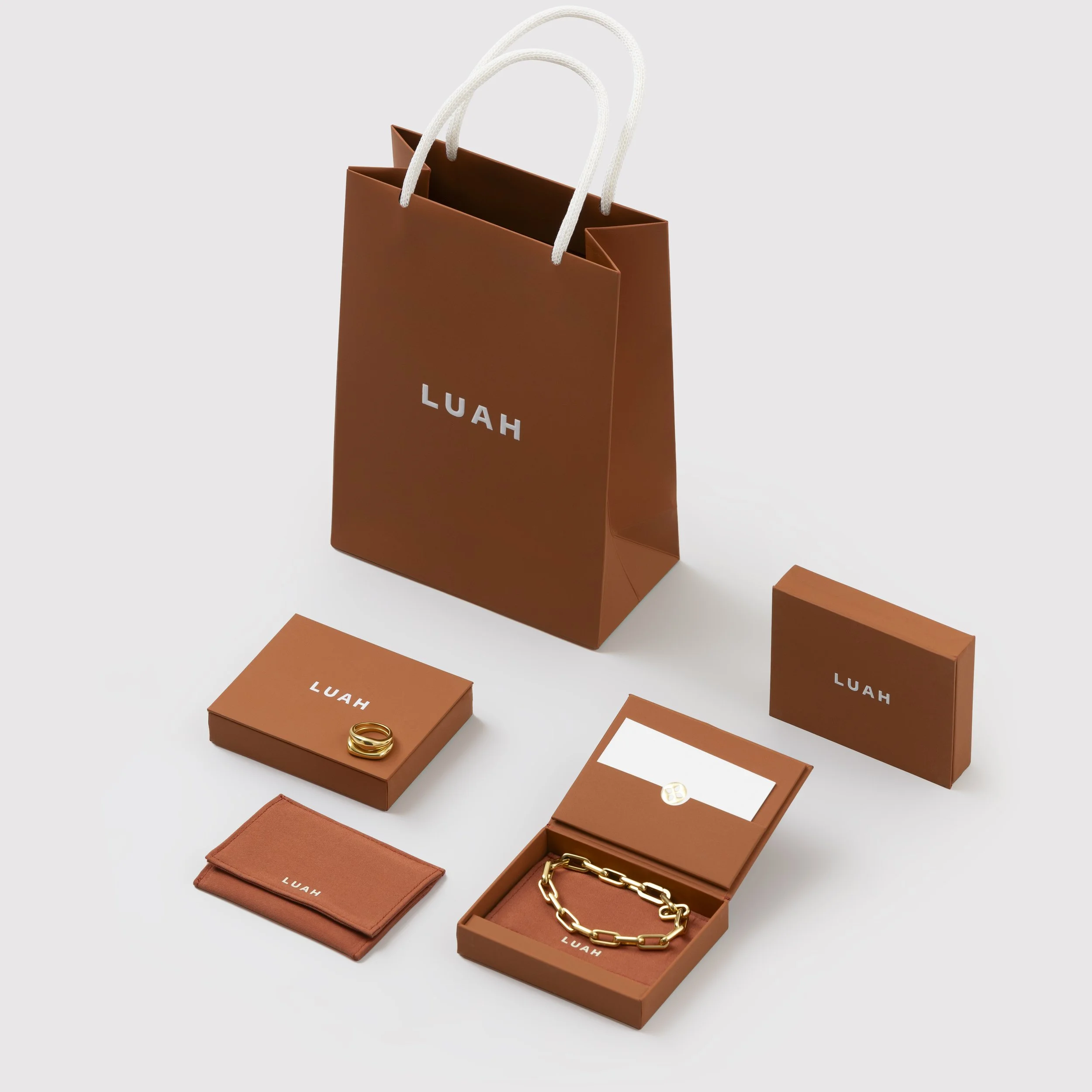 A collection of jewelry and accessories including rings, bracelets, a chain, and a cardholder all in gold, displayed alongside orange jewelry boxes labeled 'LUAH' on a white background.
