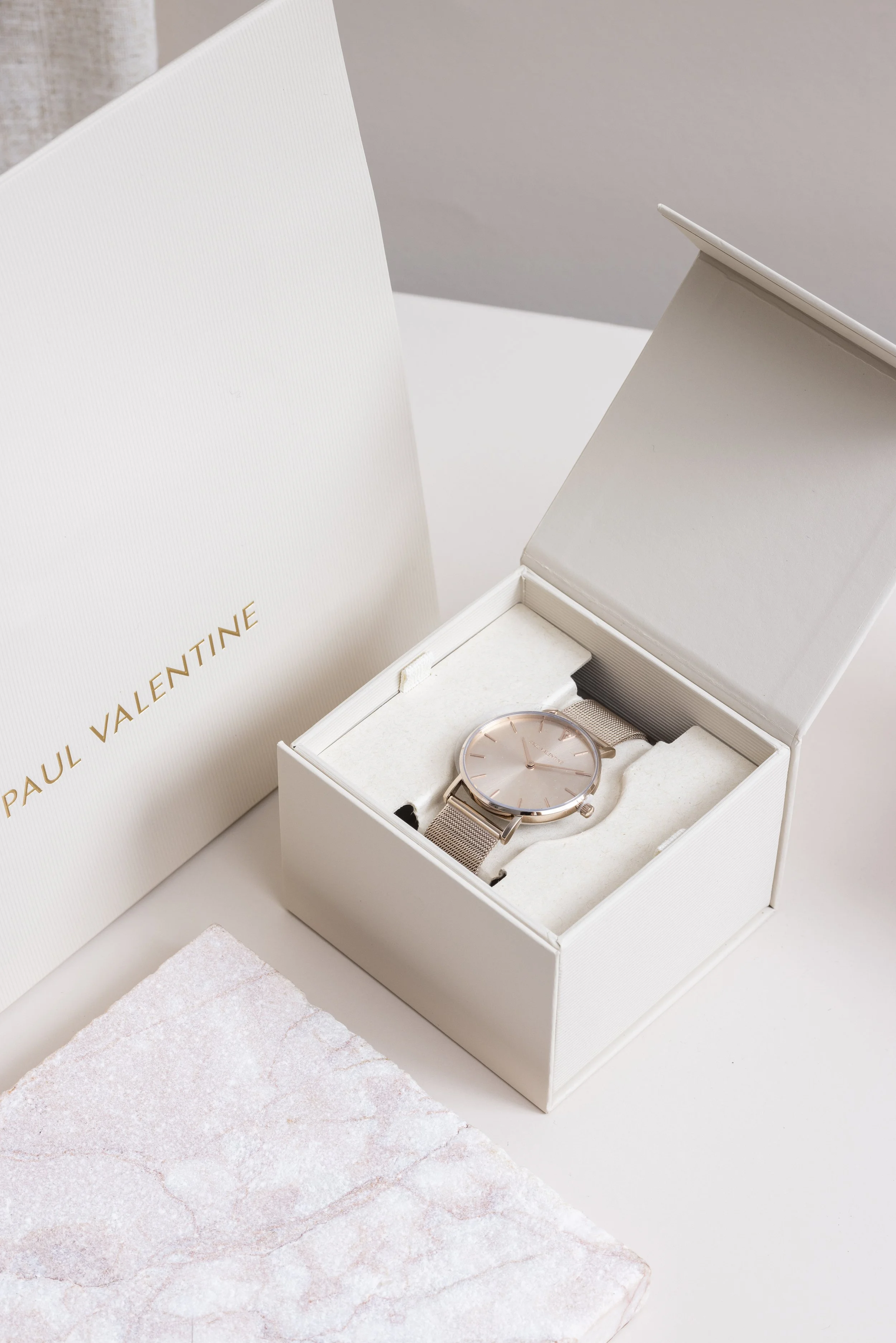 A rose gold wristwatch with a mesh band inside a white jewelry box, next to a white card with gold lettering that reads 'PAUL VALENTINE,' and a white textured card on a light surface.