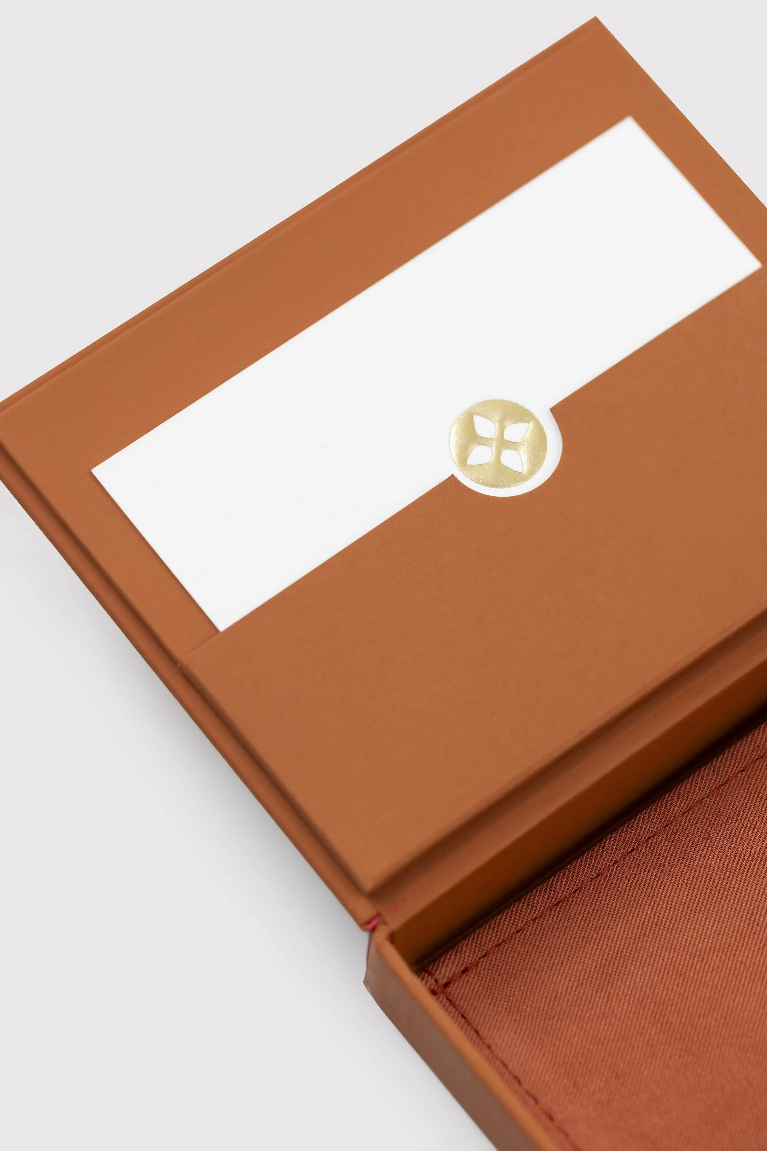 An orange jewelry box with a white label on the top and a gold decorative emblem underneath the label. The box is slightly open, revealing a padded interior lined with orange fabric.