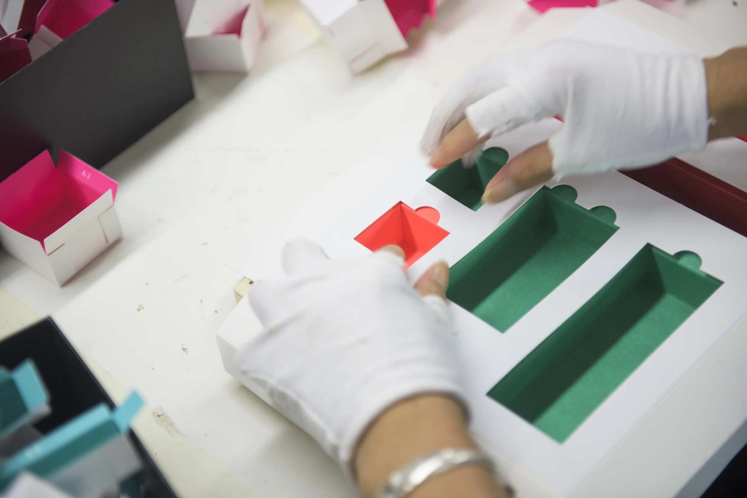 A person's hand, wearing a white glove, is placing a green game piece into a slot on a board game with colorful spaces and other game pieces.