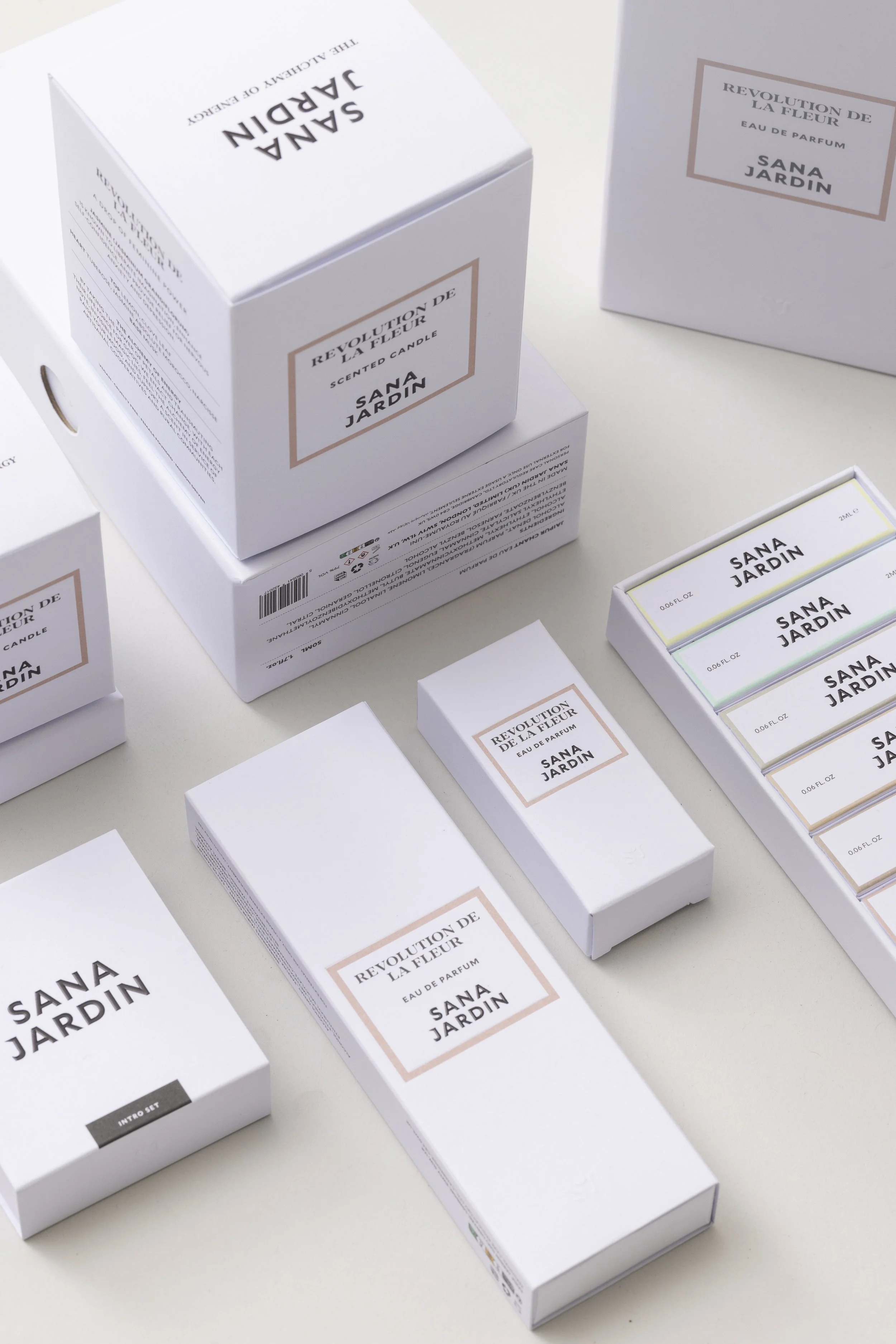 Several white boxes of Sana Jardin Revolution de la Fleur perfume are arranged on a white surface. The boxes feature minimalistic design with black and beige text, and one box is open showing small perfume sample vials.