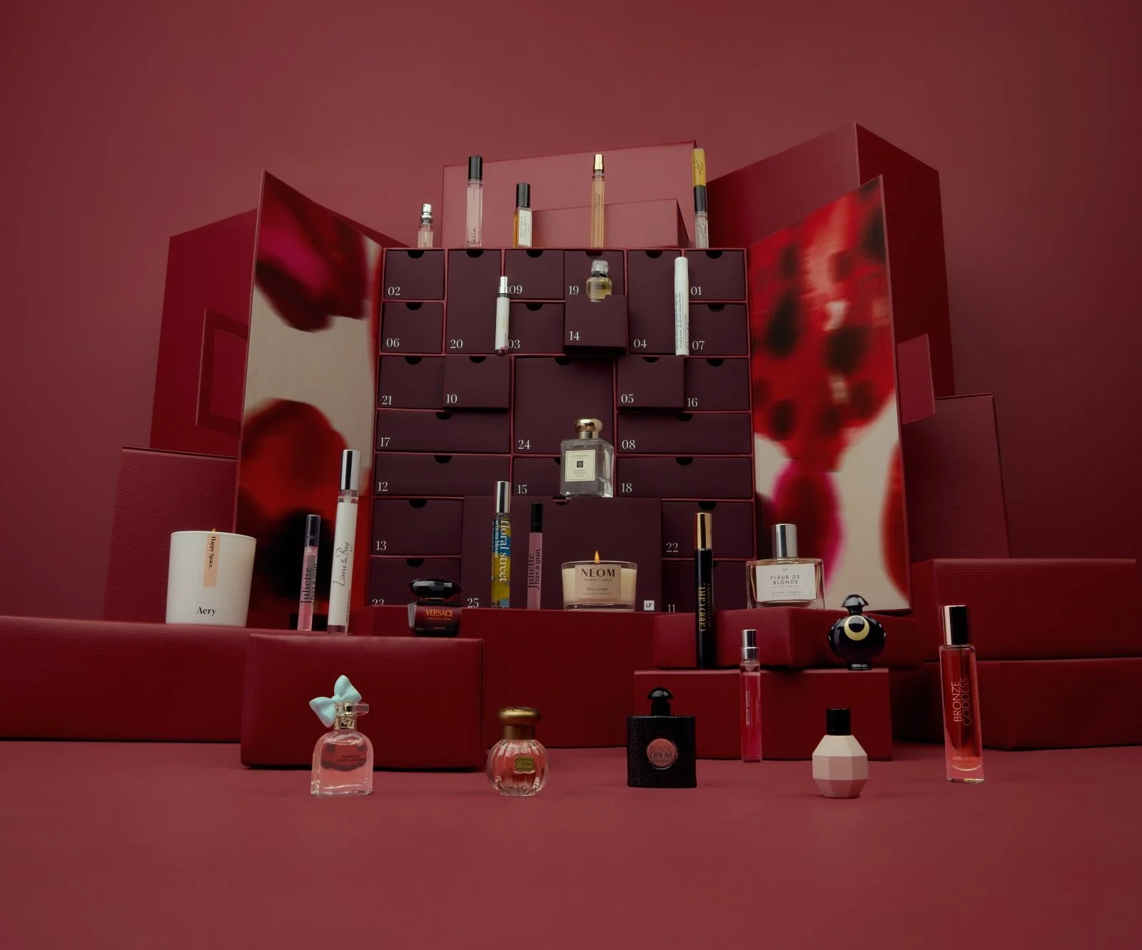 Red vanity with a dark red cabinet containing small drawers, holding various perfume bottles, candles, and beauty products. Mirrors on either side reflect the scene, with pink and red tones throughout.