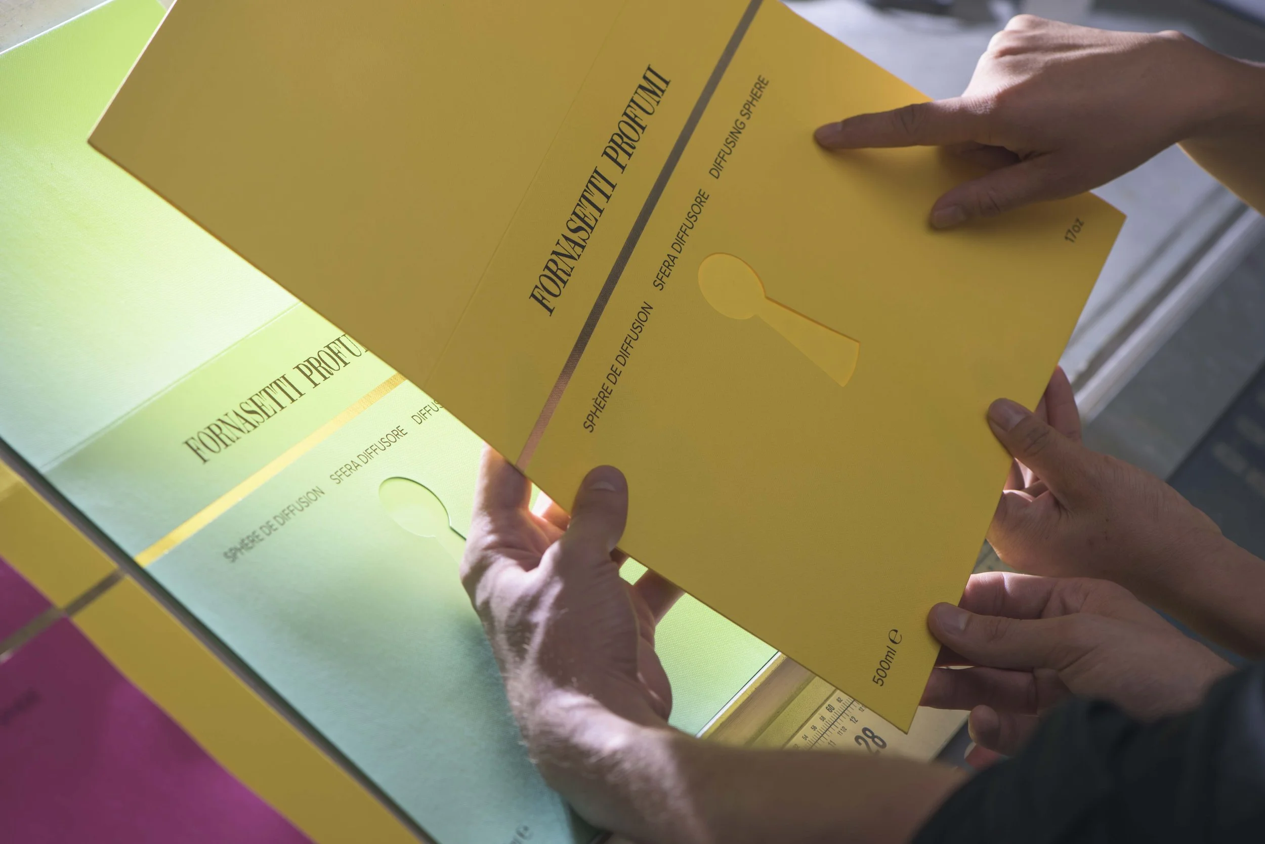 Close-up of a person's hands holding a yellow healthcare product package, labeled 'Fornasetti Profumi,' with a keyhole graphic and text indicating it is a diffuser sphere with 500ml volume; another package is visible in the background.