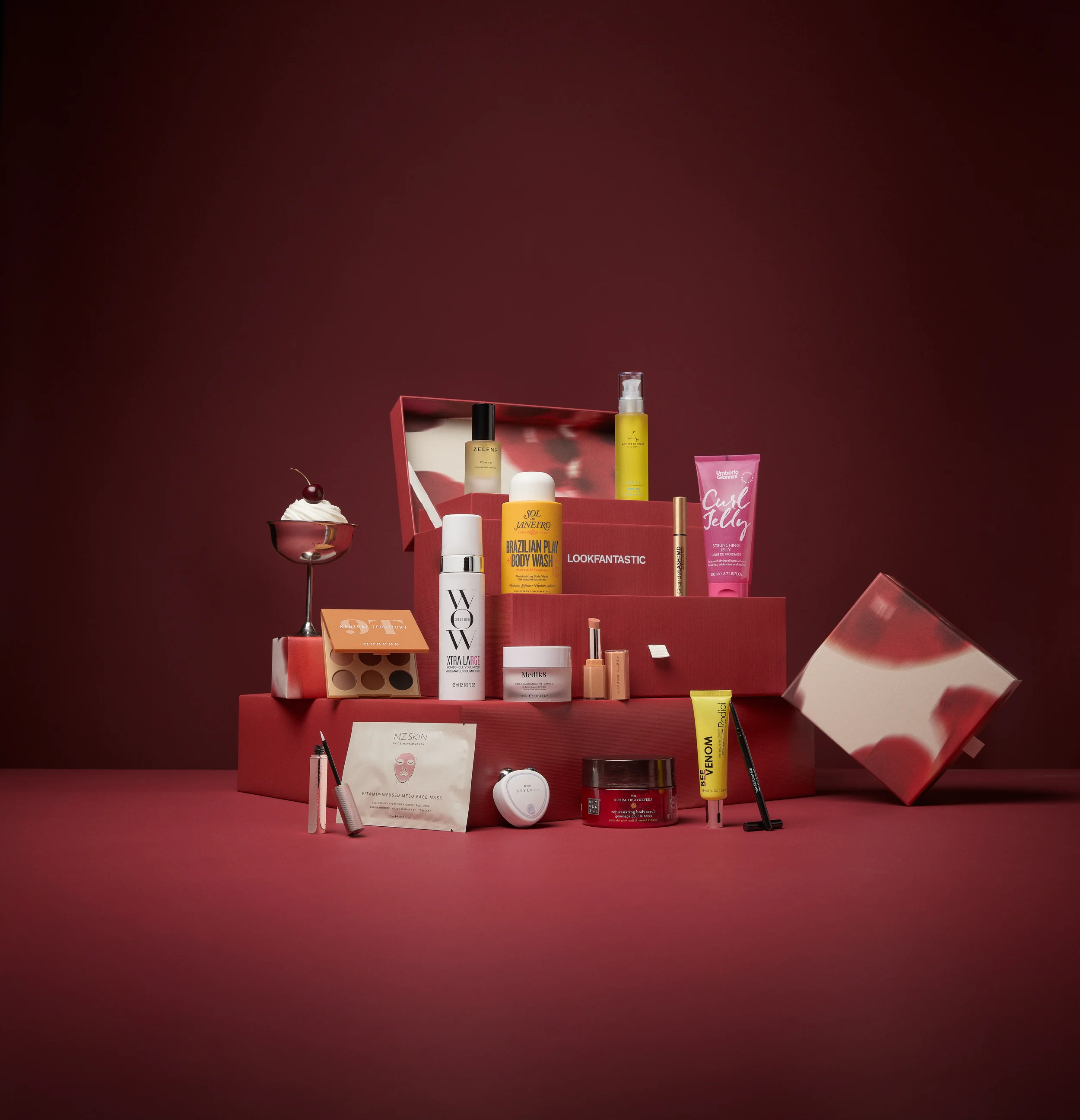 A pink gift box containing skincare and beauty products, including bottles, tubes, and a sheet mask, with a glass of whipped cream dessert topped with a cherry to the left.
