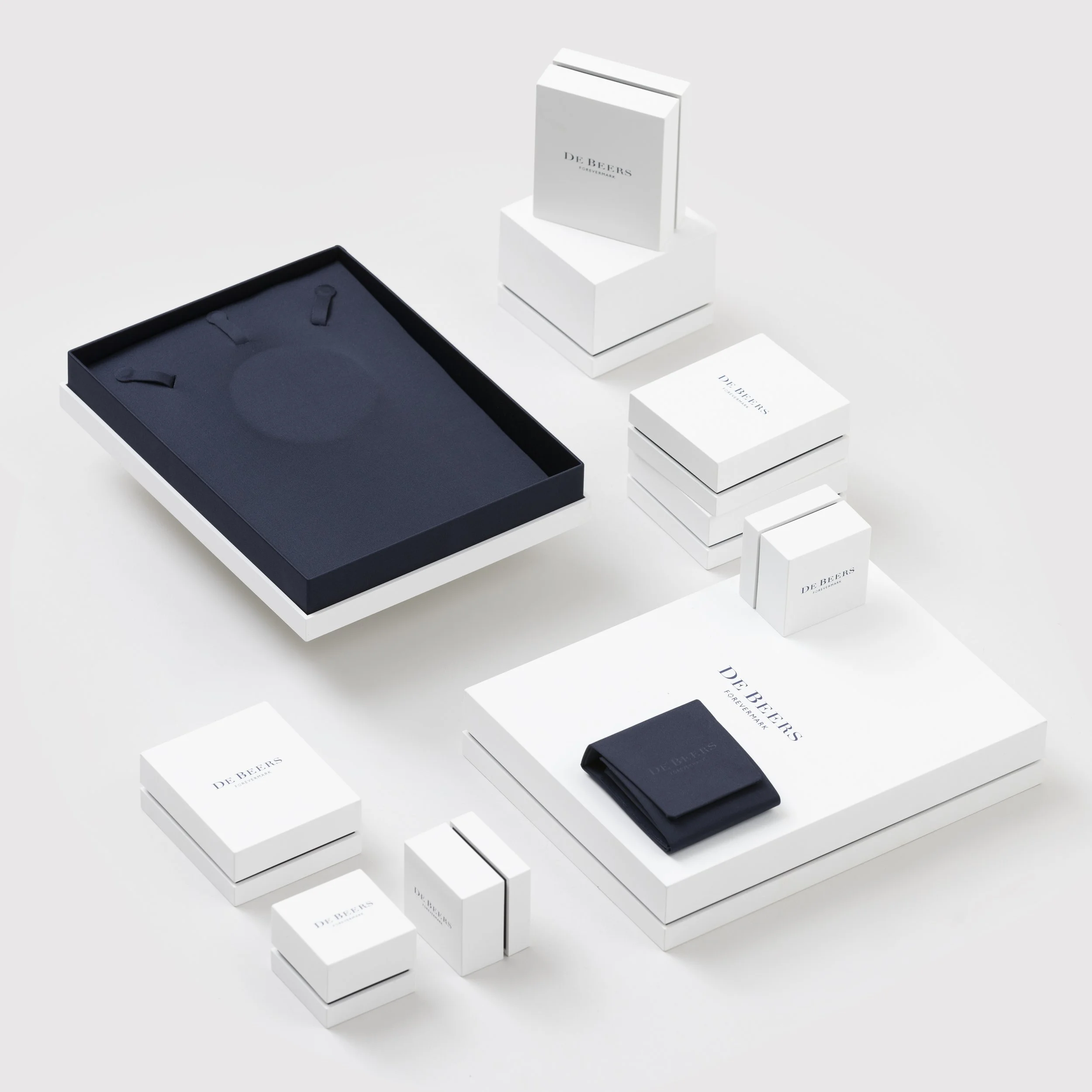 A collection of white boxes and packaging with the logo 'DE BEERS' on them, arranged on a white surface. Some boxes are stacked, while others are laid flat or slightly opened, showcasing the minimalist design.