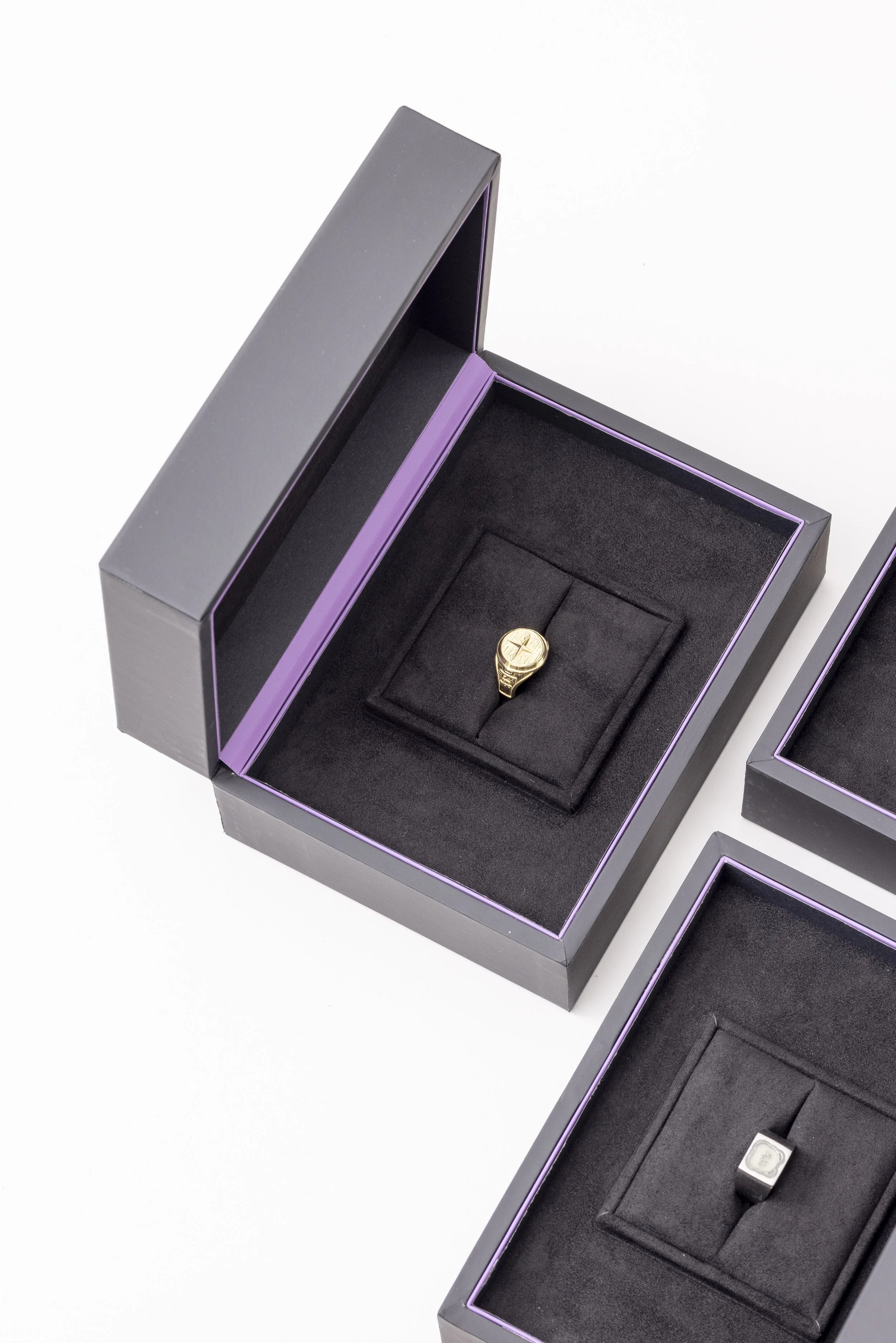 Close-up of two black jewelry boxes with purple edges, one open revealing a gold watch inside, the other closed, on a white background.