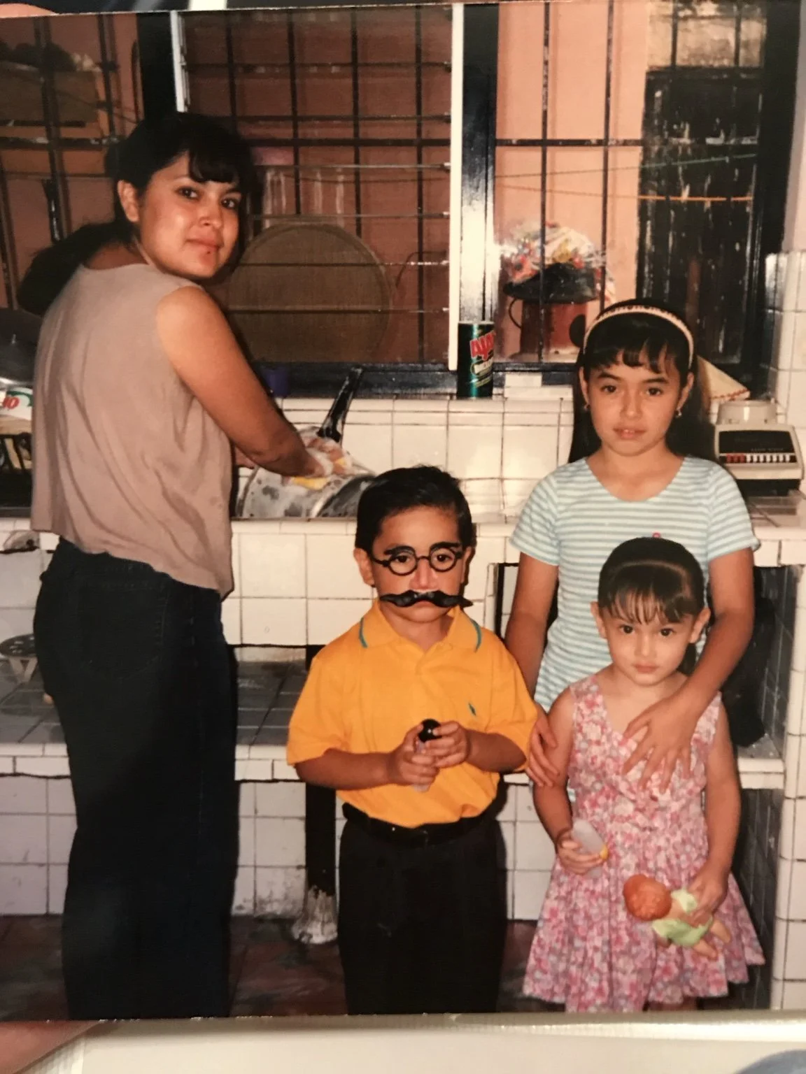 A woman and three children in a kitchen, with the woman washing dishes at the sink and the children posing for the photo. The children are a boy with glasses and a fake mustache, a girl with a headband, and a girl holding ice cream cones.