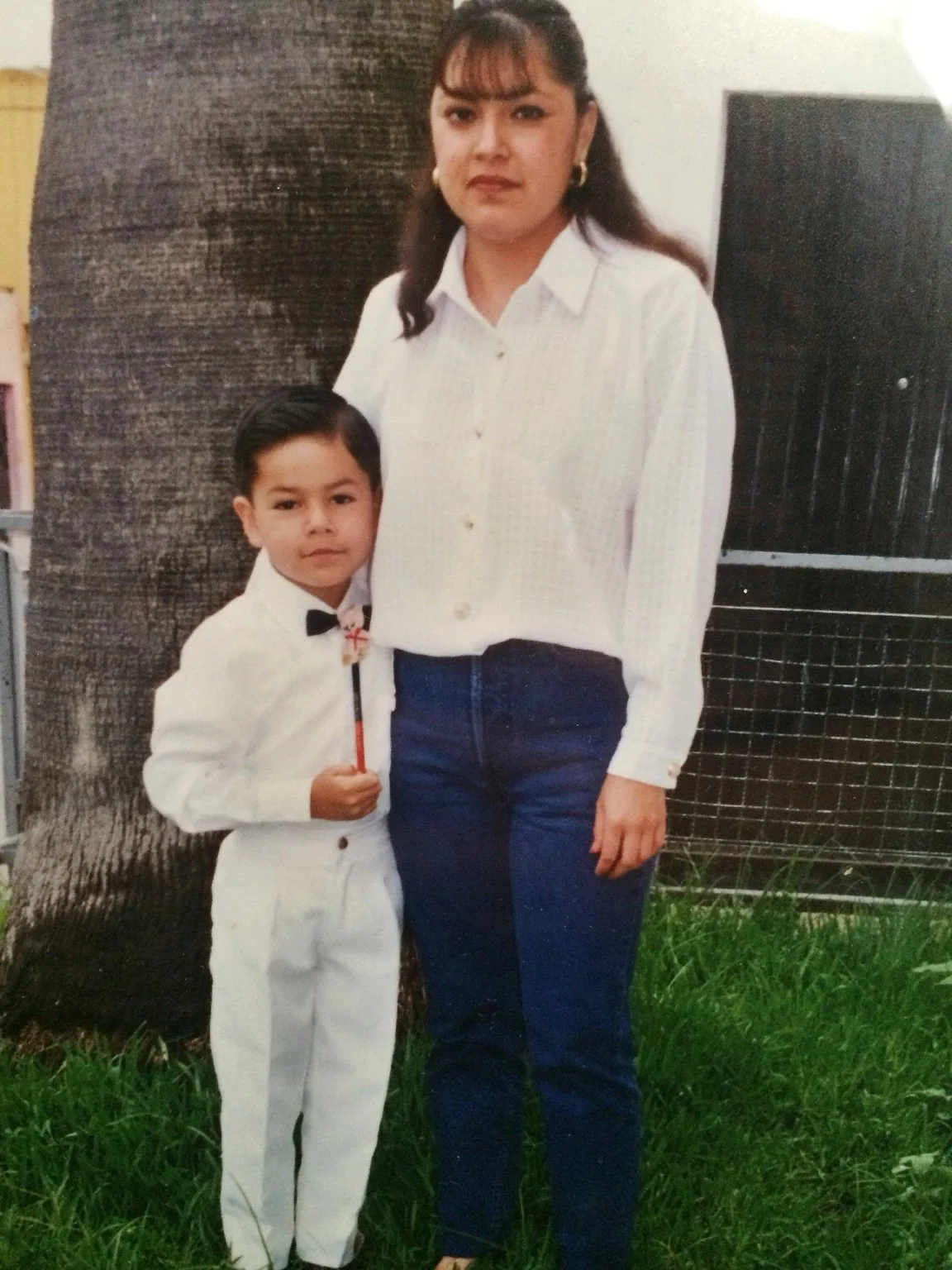 A woman and a young boy standing outdoors in front of a large tree. The woman is wearing a white blouse and jeans, with dark hair and earrings. The boy is wearing a white suit with a bow tie and is holding a thin stick or pointer.