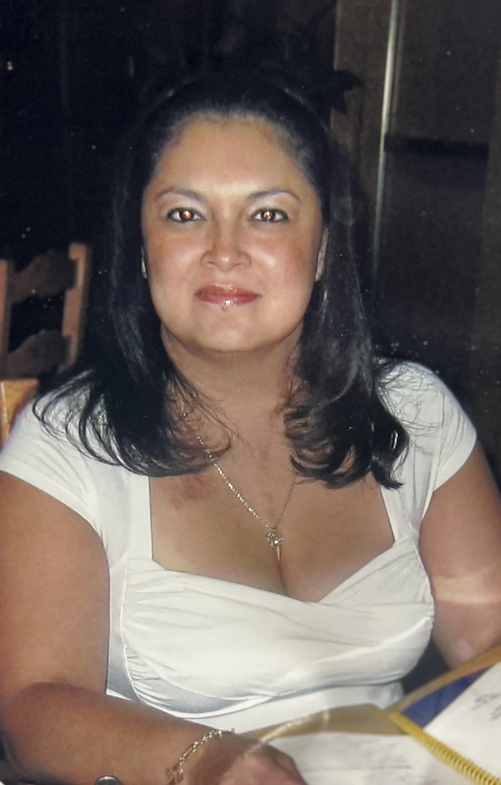 A woman with long black hair, wearing a white top, gold jewelry, and makeup, smiling at the camera, seated indoors.