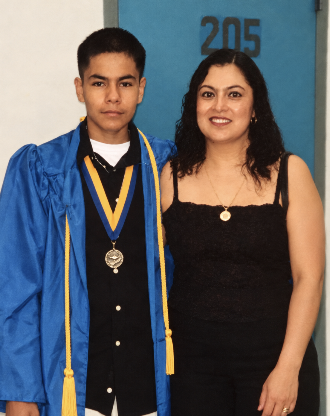 A young man in a blue graduation gown and medals stands with a woman in a black dress, both smiling, in front of a blue door with the number 205.