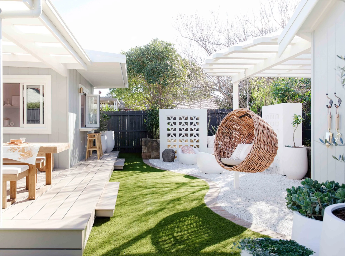 A backyard patio with a small lawn and a sitting area under a white pergola, featuring a woven hanging chair, potted plants, and outdoor furniture in a bright, modern style.