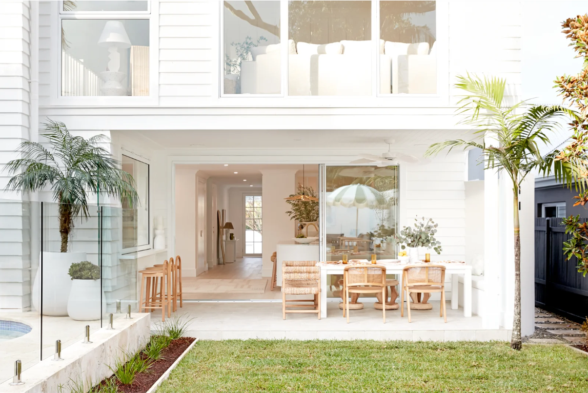 The image features the backyard patio area of a modern white house with large glass sliding doors. There is a dining table with chairs outside, set for a meal, with lush green plants and a grassy lawn in the foreground. The interior visible through the sliding door shows a bright, minimalistic living space with a white couch, indoor plants, and natural light.