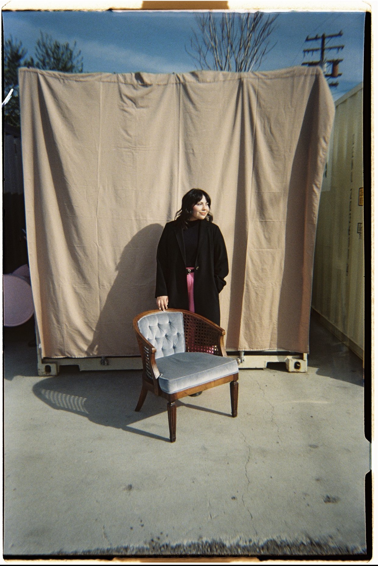 A woman with dark hair standing in front of a beige curtain backdrop outdoors, with a chair with a light-colored cushion and wooden frame in front of her and a blue sky with some trees and power lines in the background.