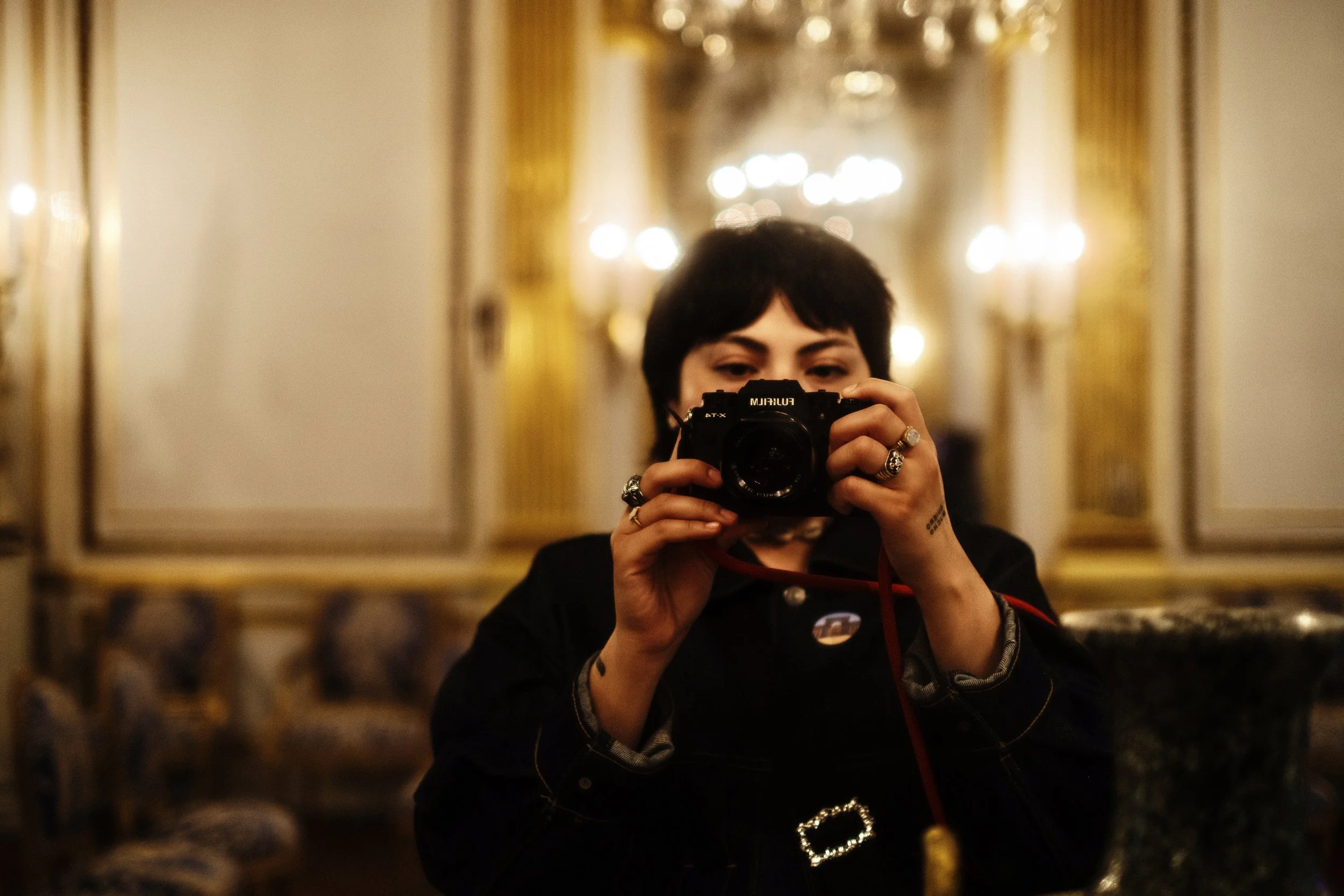 Person with short black hair taking a selfie in front of a mirror, surrounded by warm yellow lighting in an elegant room.