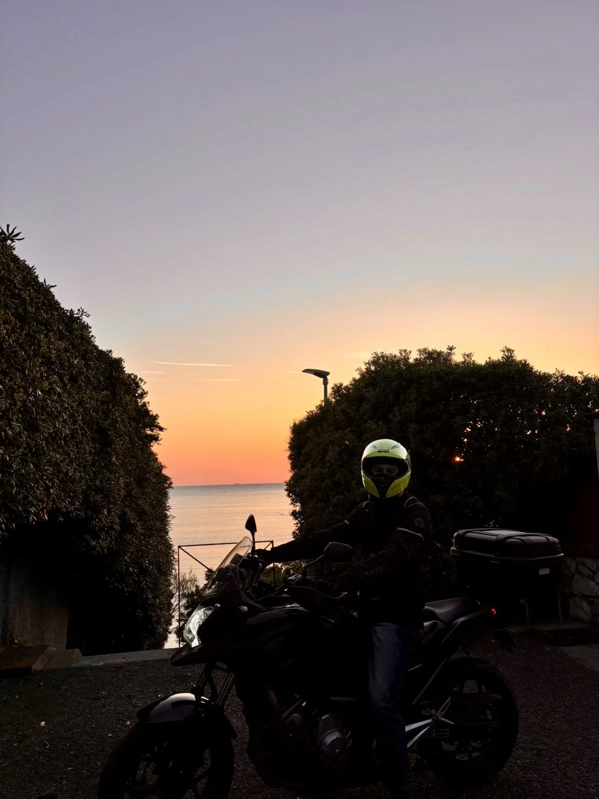 Un motociclista con casco giallo e nero, vestito di nero, su una moto davanti a un tramonto sul mare, tra due siepi.