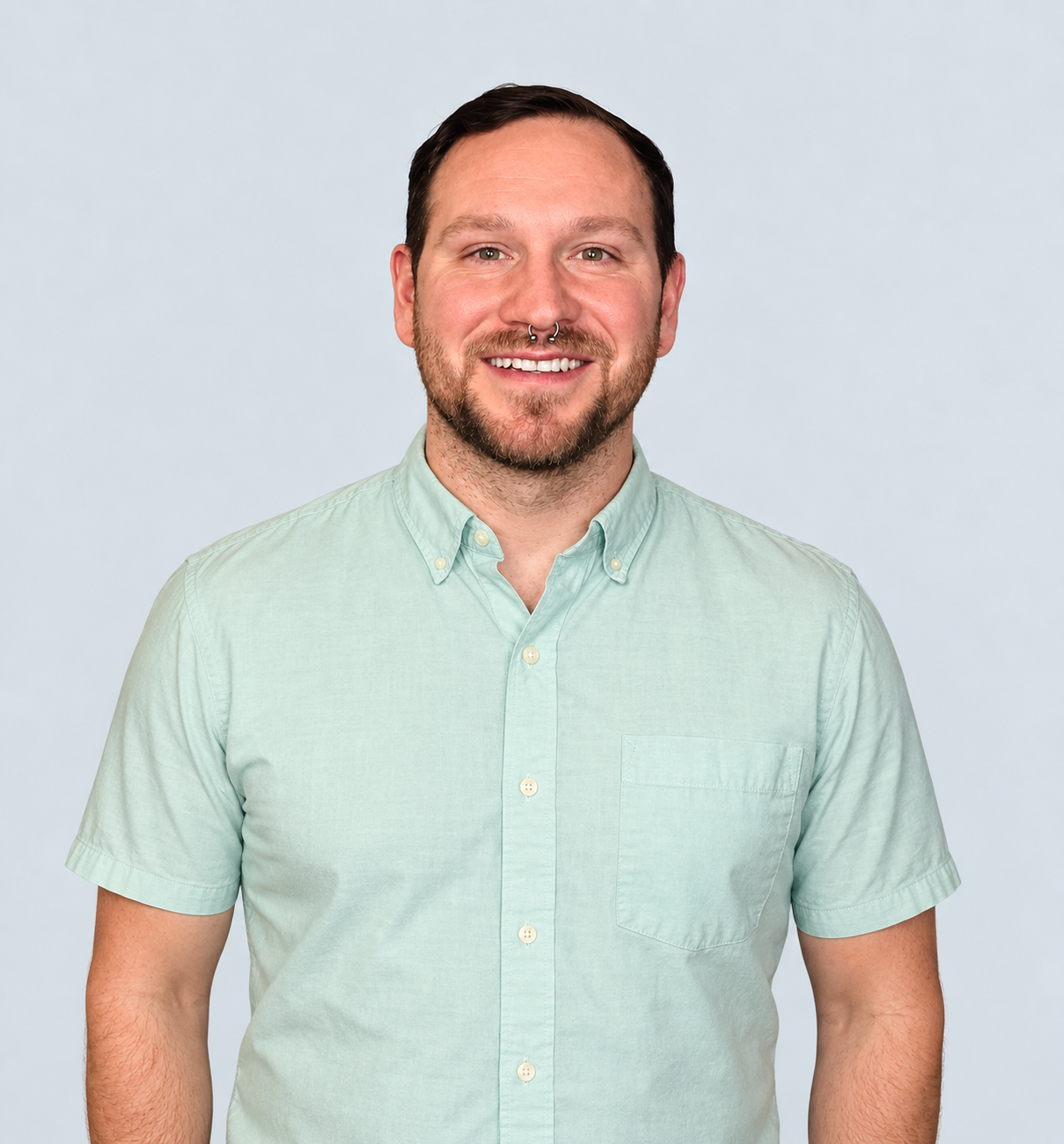Headshot of a man with short dark hair, beard, and septum piercing, wearing a light green button-up shirt, smiling against a light gray background.