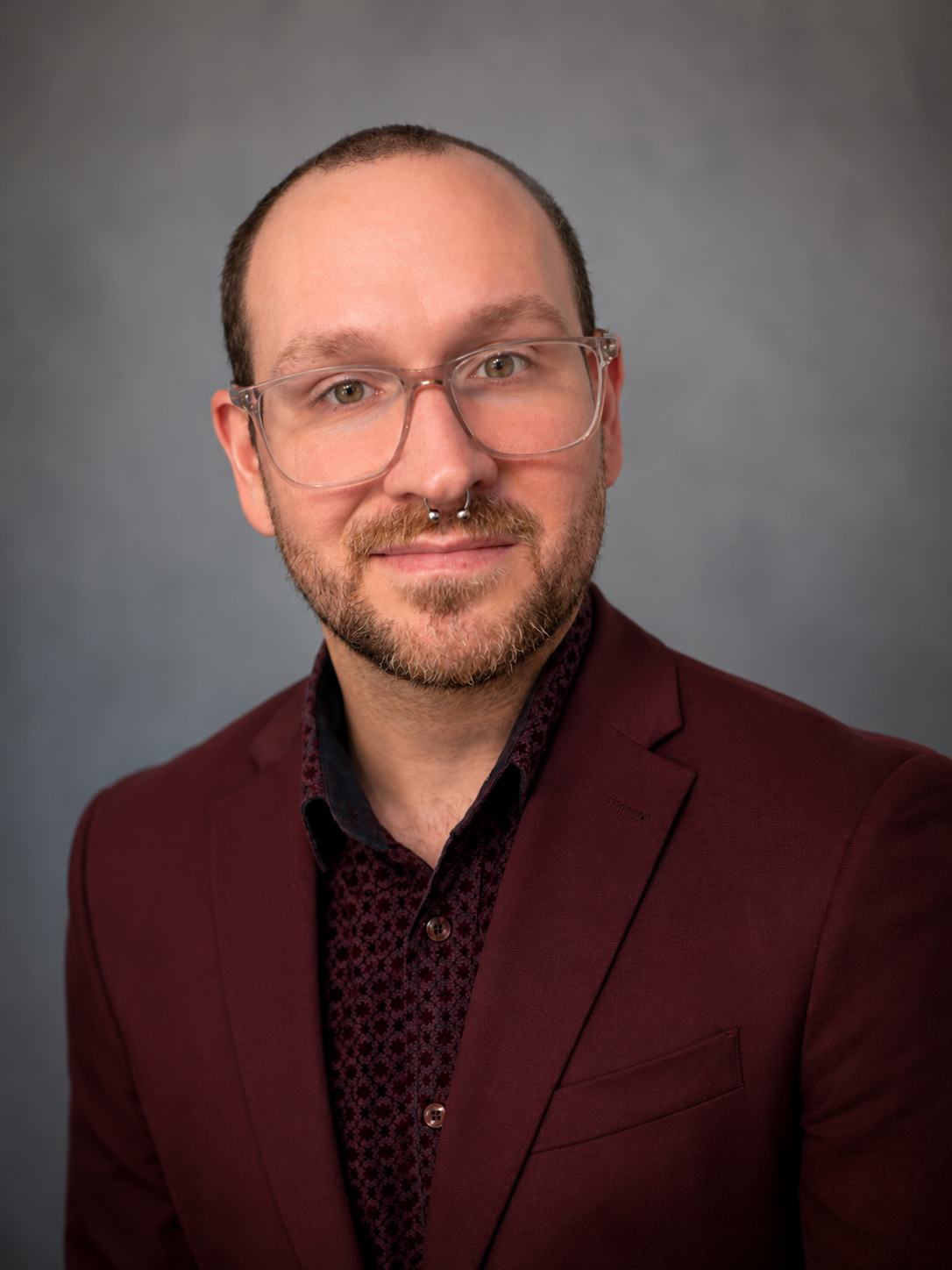 Headshot of a man with glasses, a septum piercing, a beard, wearing a burgundy blazer and patterned shirt, against a gray background.