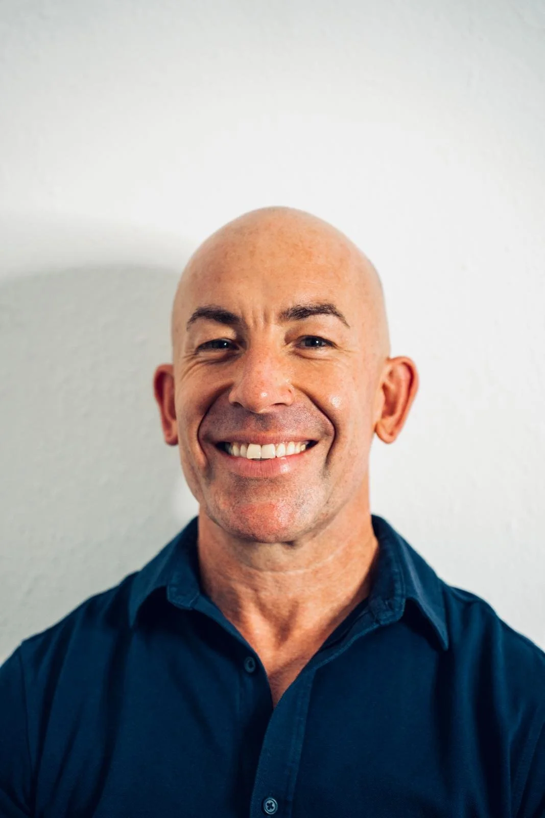 A smiling bald man wearing a dark blue shirt, standing against a plain white wall.