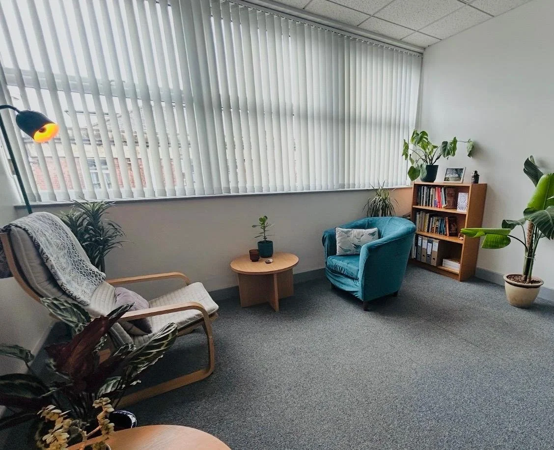 Office space with two chairs, a small wooden table, a bookshelf, and numerous potted plants near large window with vertical blinds.