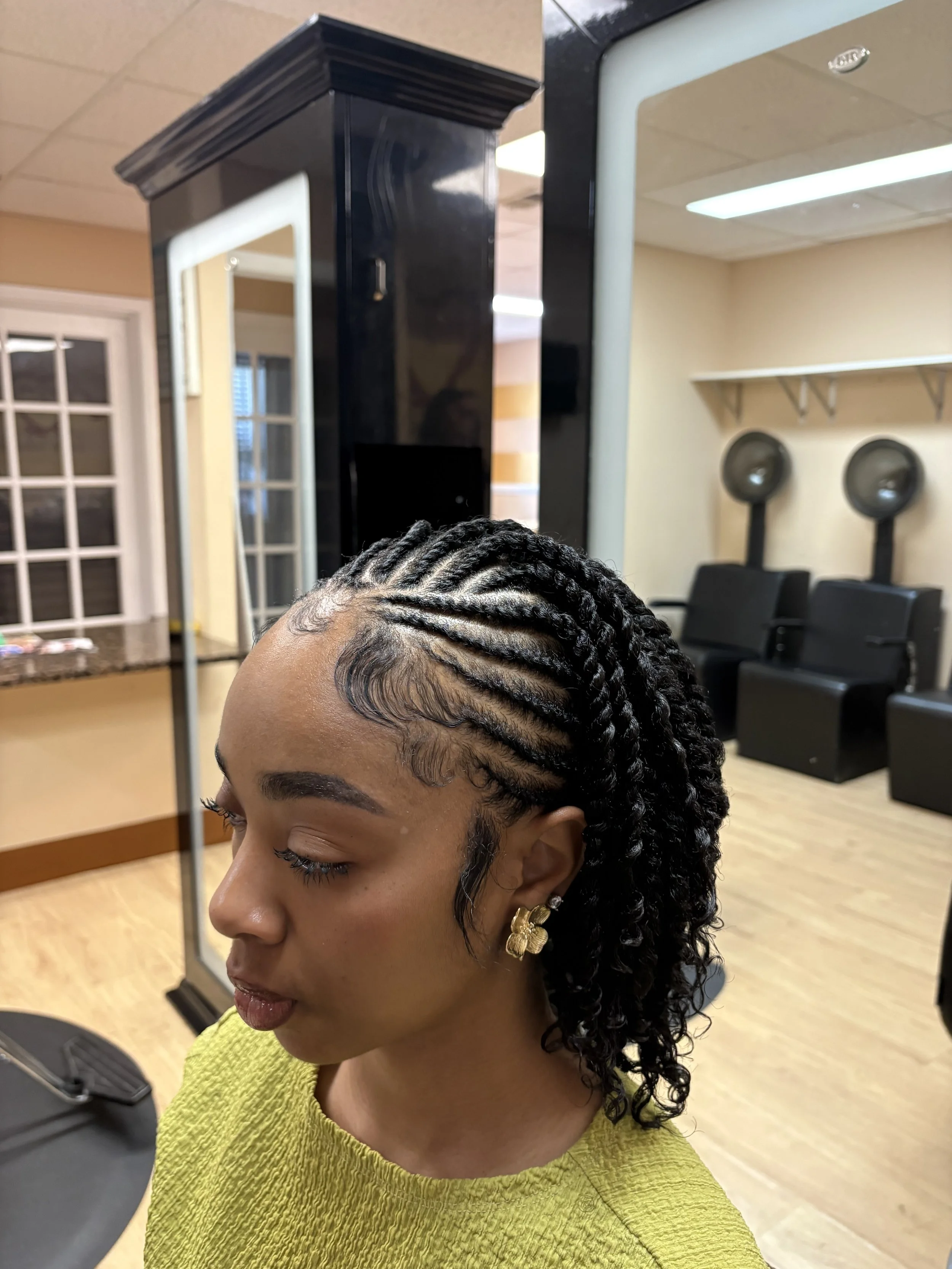 A woman with braided hair and gold earrings is sitting in a salon or barbershop, with hair care chairs and mirrors visible in the background.