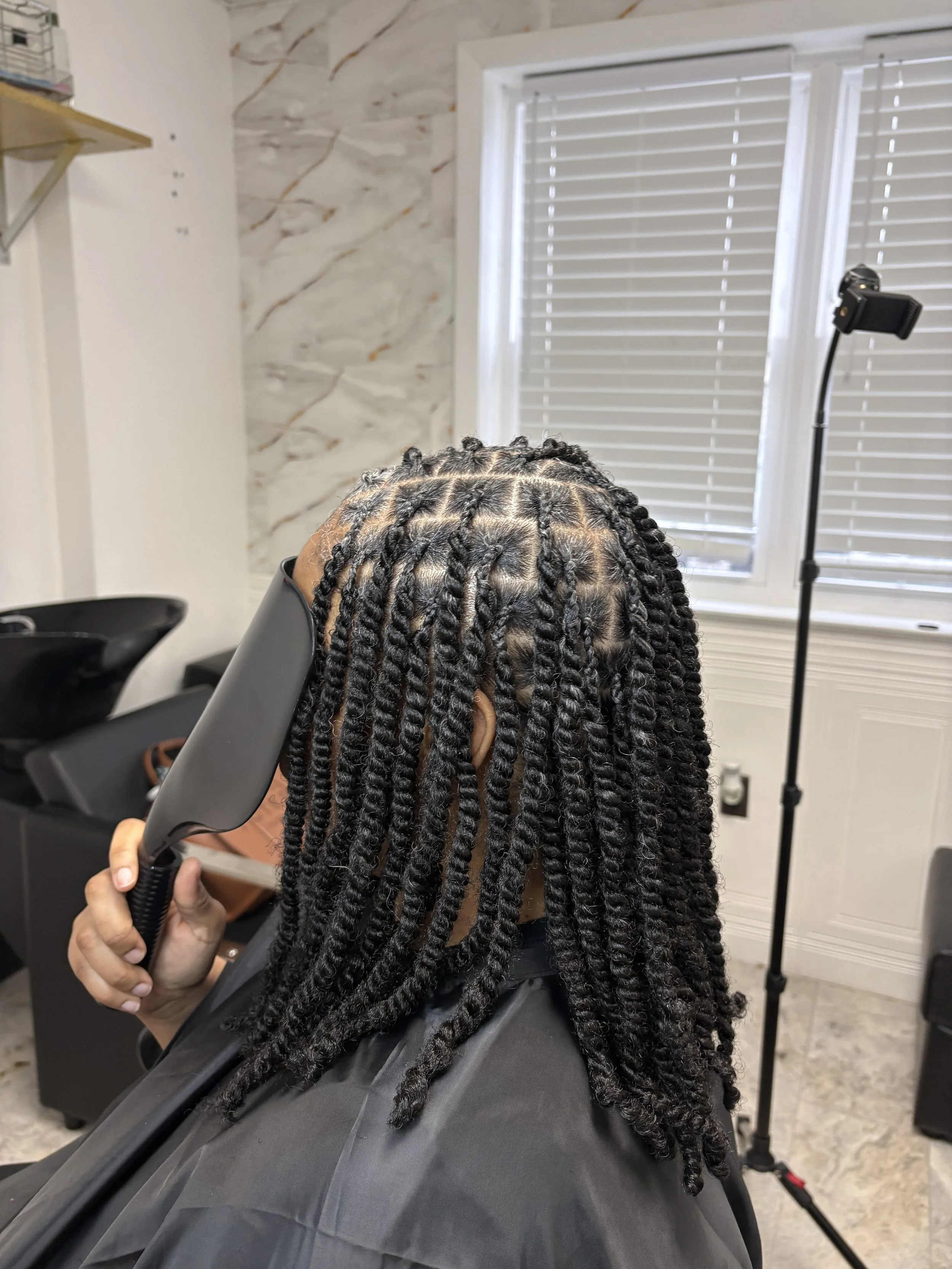 Person with braided hair sitting in a salon chair, getting their hair shaped with a clipper, in a hair salon with a window and a stand with a camera or phone mounted on it.