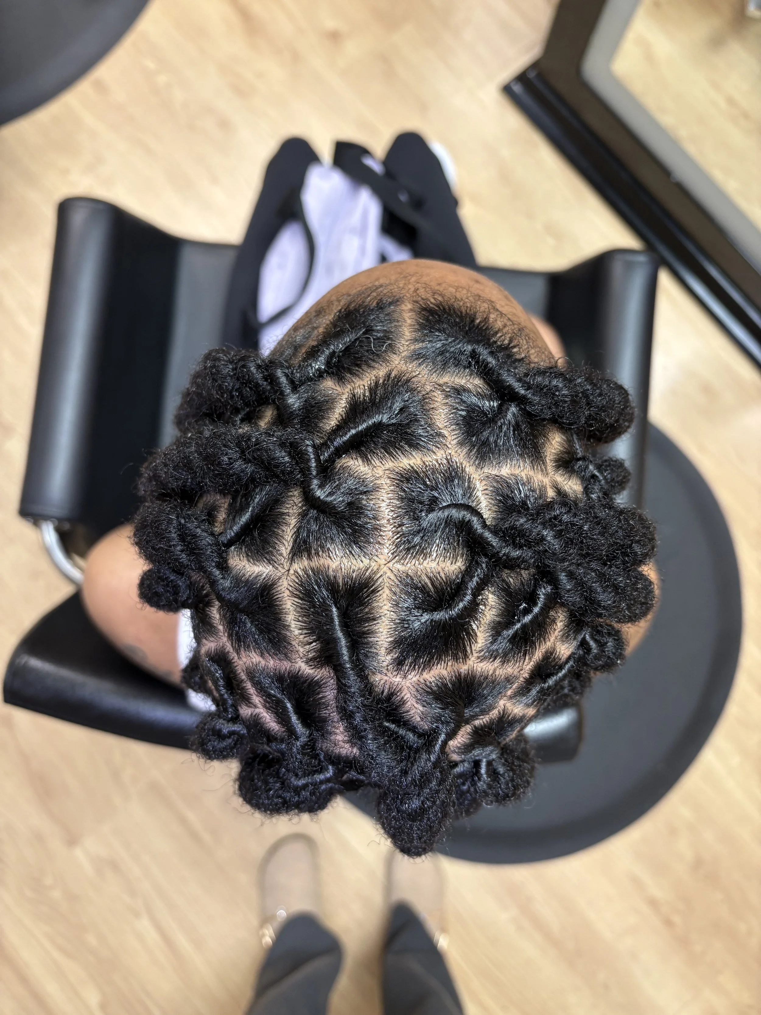 Top-down view of a person with intricately styled, coiled braids, sitting on a black chair, with a light-colored wooden floor beneath.