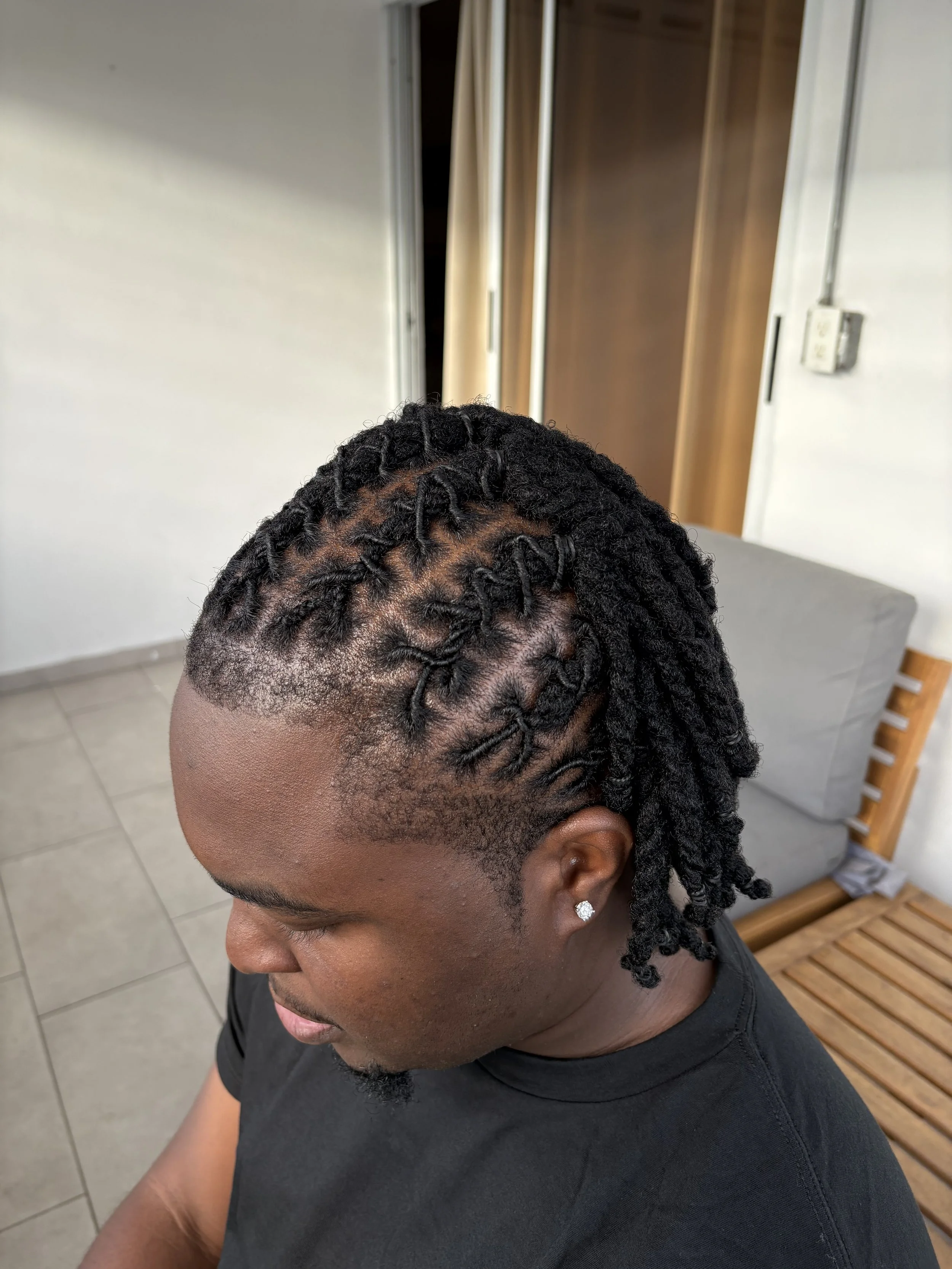 Close-up of a woman's head showing styled dreadlocks, with her face partially visible, sitting indoors near a wooden chair and a gray couch.