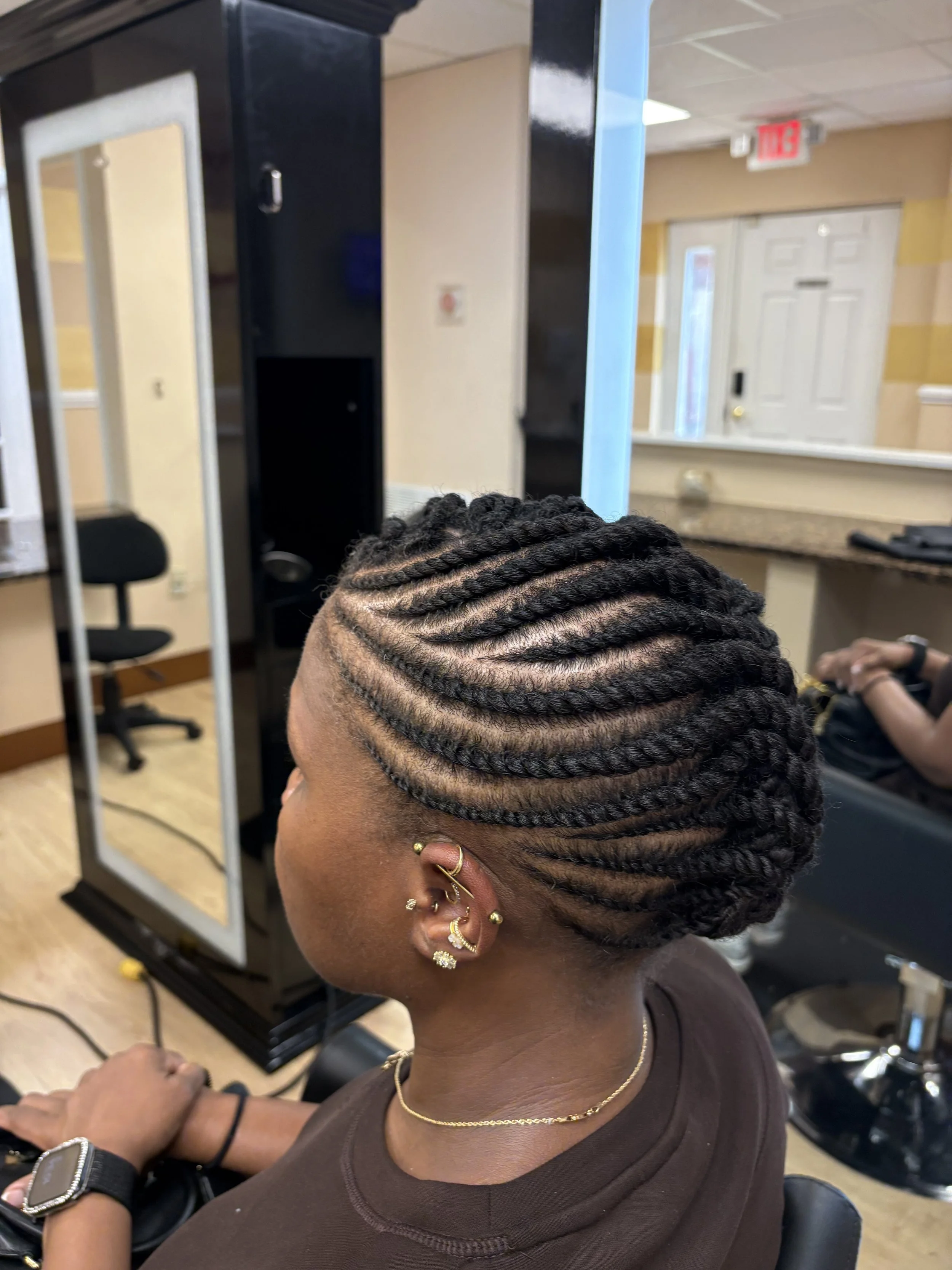 Close-up of a person with neatly braided hair, showing multiple cornrow braids running from the front to the back. The individual is sitting in a salon chair, wearing earrings, a gold necklace, and a black wristwatch, with a salon environment in the background.