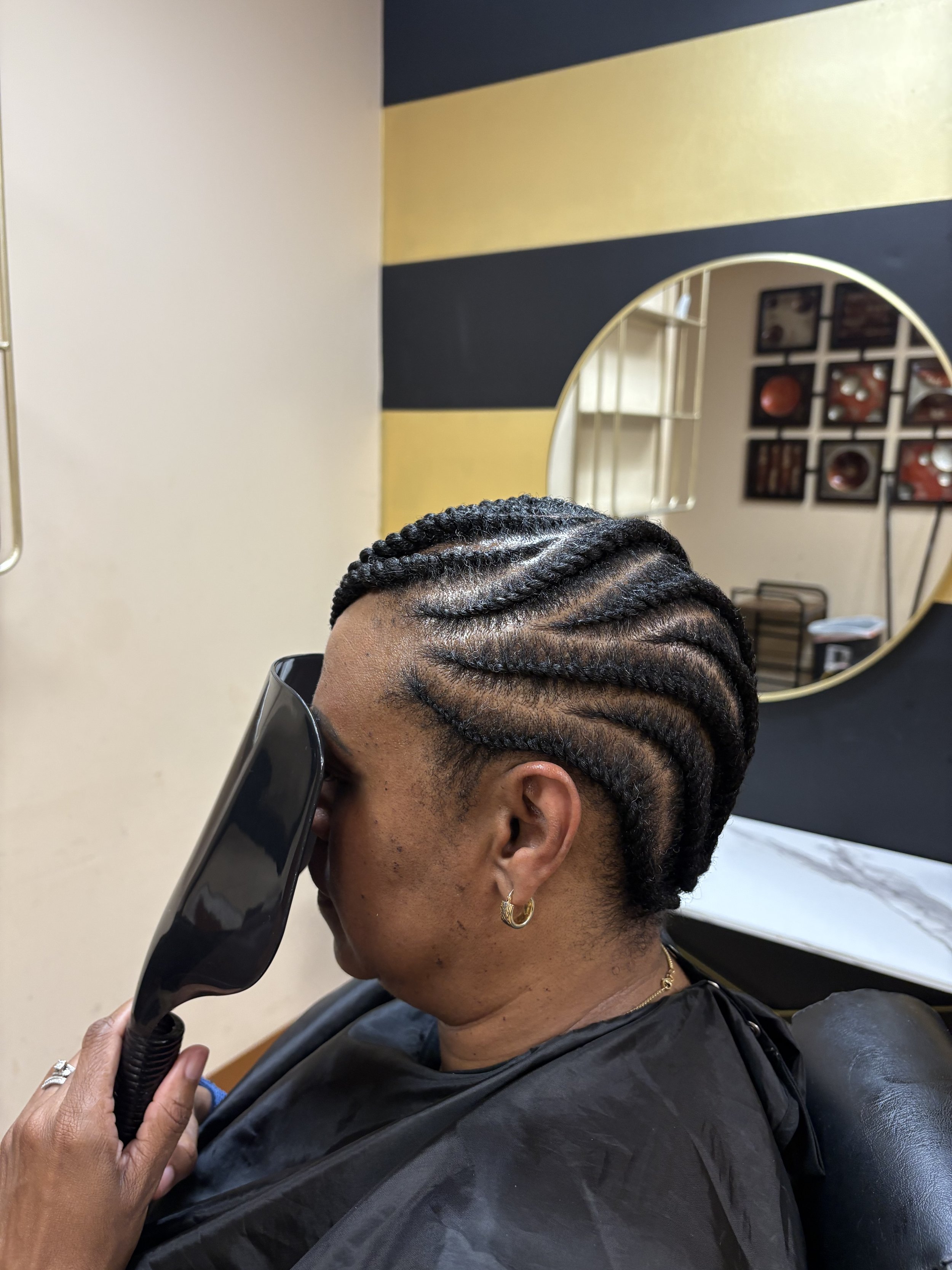 A person with braided hair and gold earrings sitting in a salon, while a stylist uses a handheld vacuum to clean their hair.