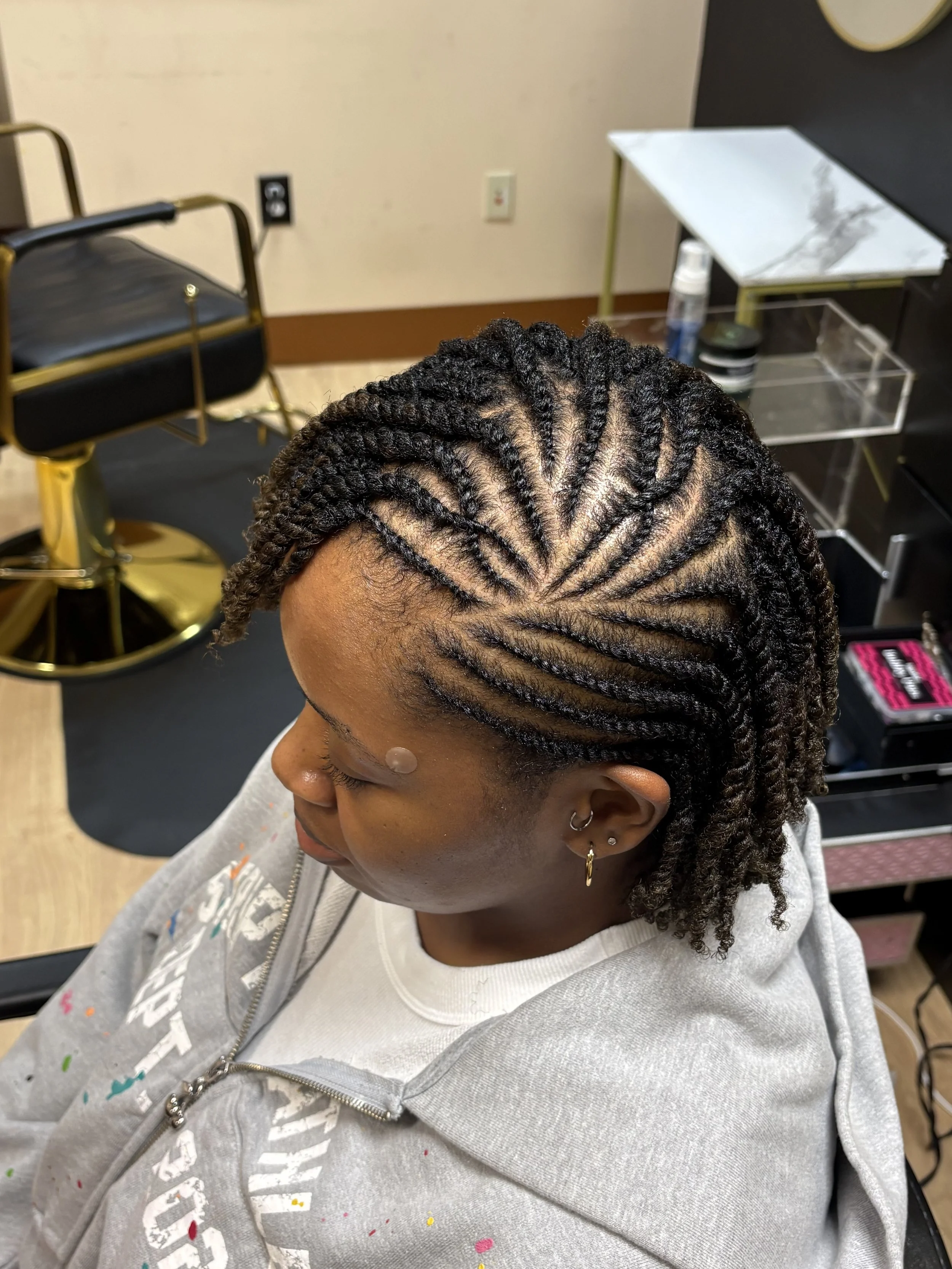 A woman with braided hairstyle sitting in a salon chair, with a hairdresser's station and salon equipment visible in the background.