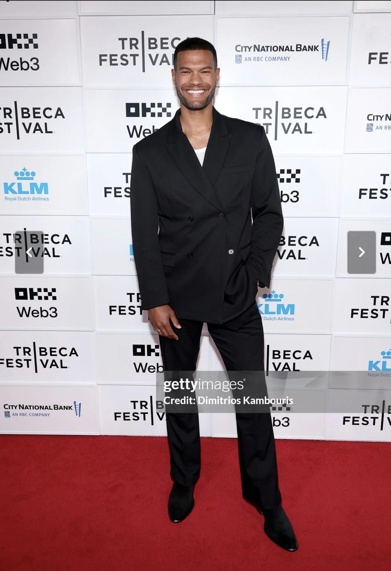 A man dressed in a black suit and white shirt standing on a red carpet at Tribeca Festival, smiling at the camera with a backdrop featuring festival and sponsor logos.