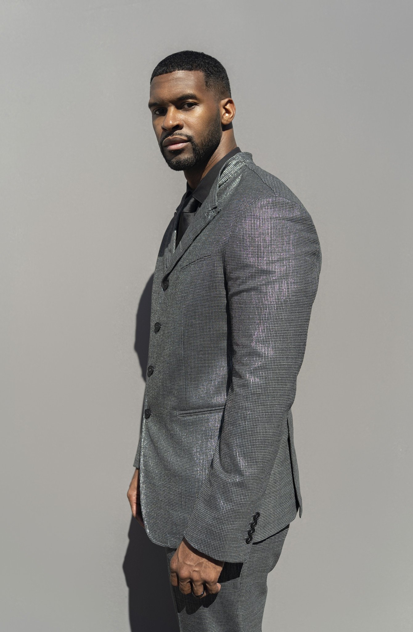 A man with a beard and short haircut wearing a gray plaid suit, black shirt, and black shoes standing against a plain gray background.