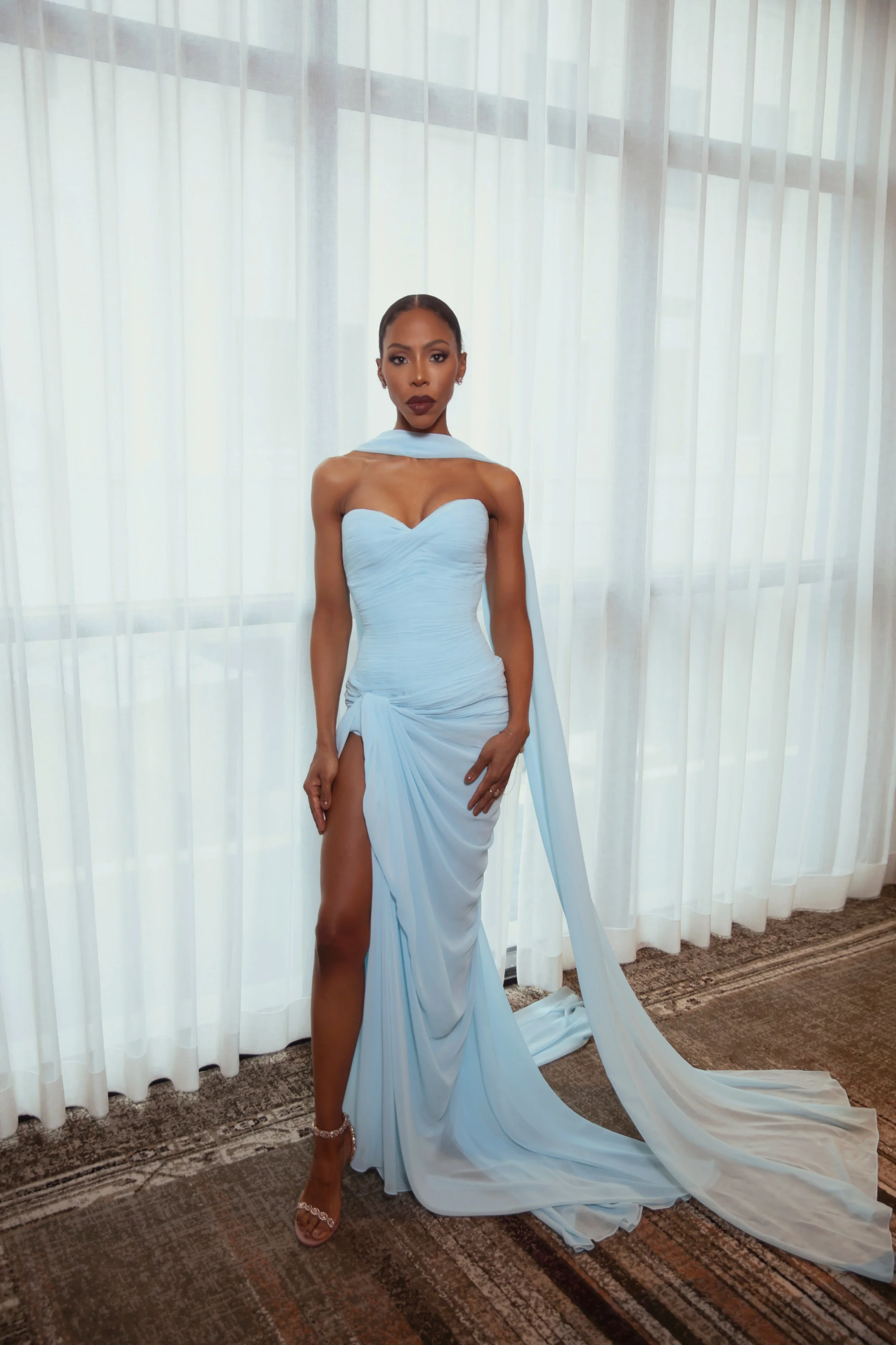 A woman wearing a light blue gown with a high slit, standing in front of sheer white curtains, with her right leg exposed, and her hair styled in a sleek bun.