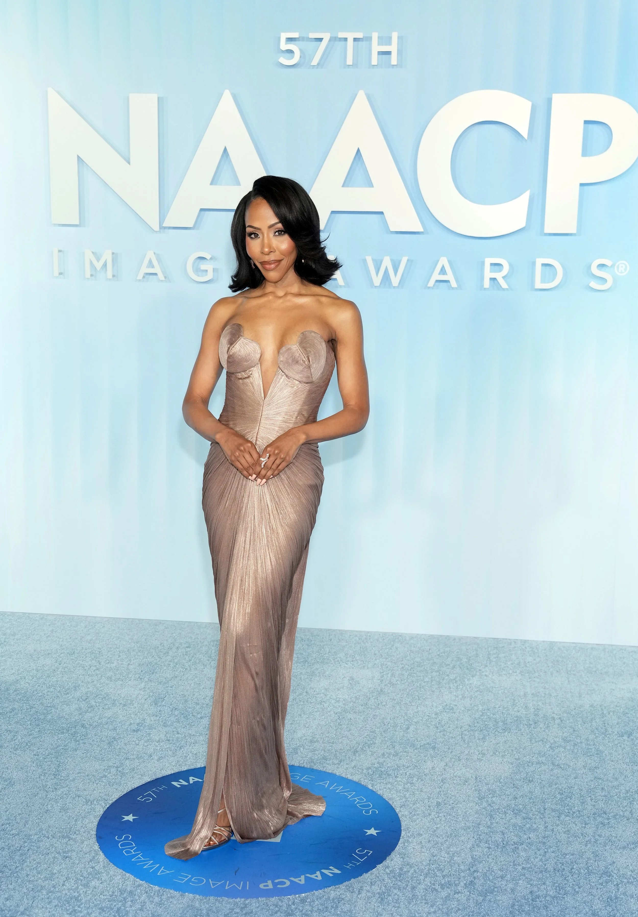 A woman in a shimmering gown standing on a blue circular platform at the 57th NAACP Image Awards, with a light blue backdrop featuring the event logo.