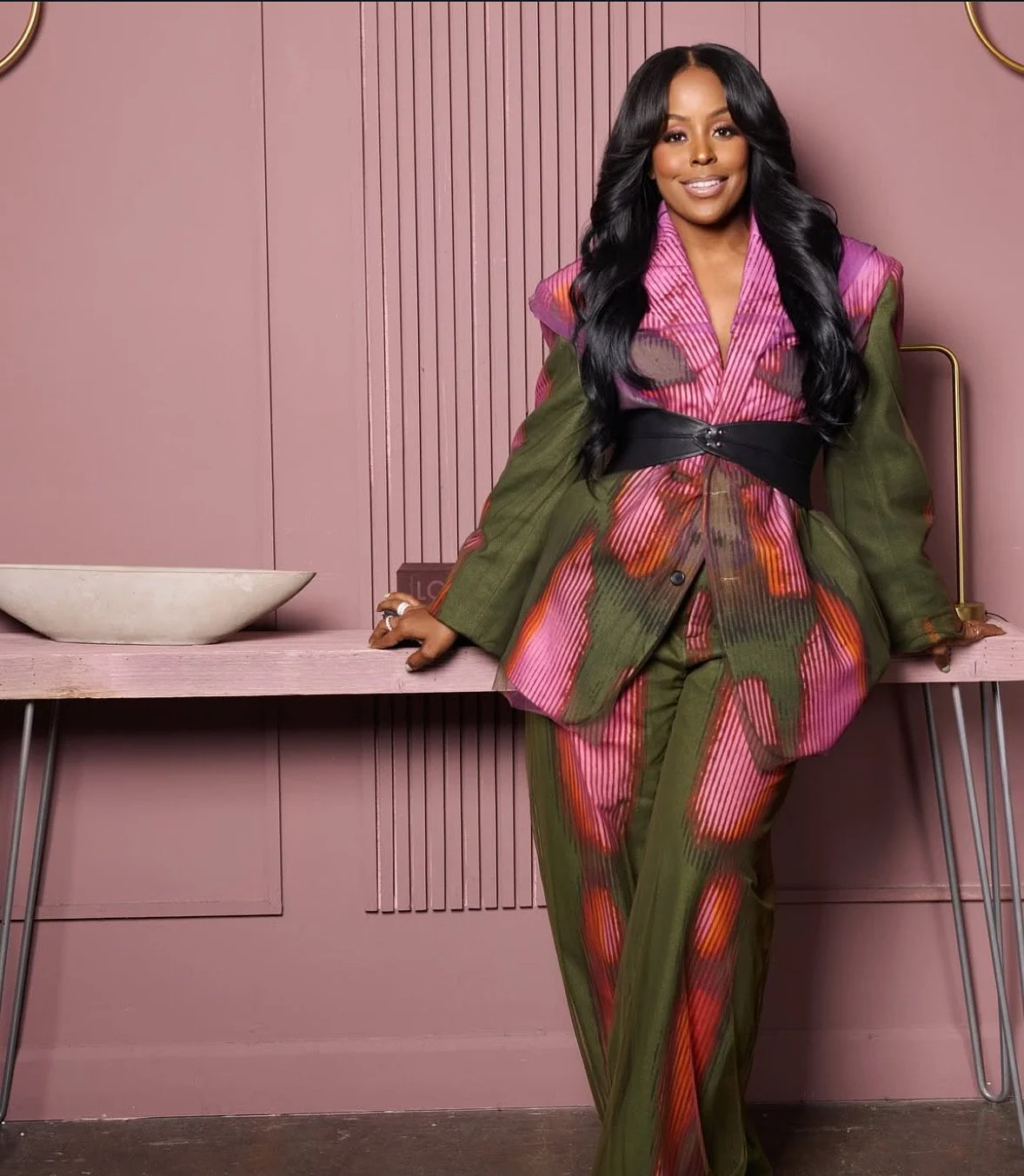 A woman dressed in a colorful floral-patterned suit, standing against a pink wall with vertical paneling, leaning on a pink table with a white bowl and gold lamp in the background.