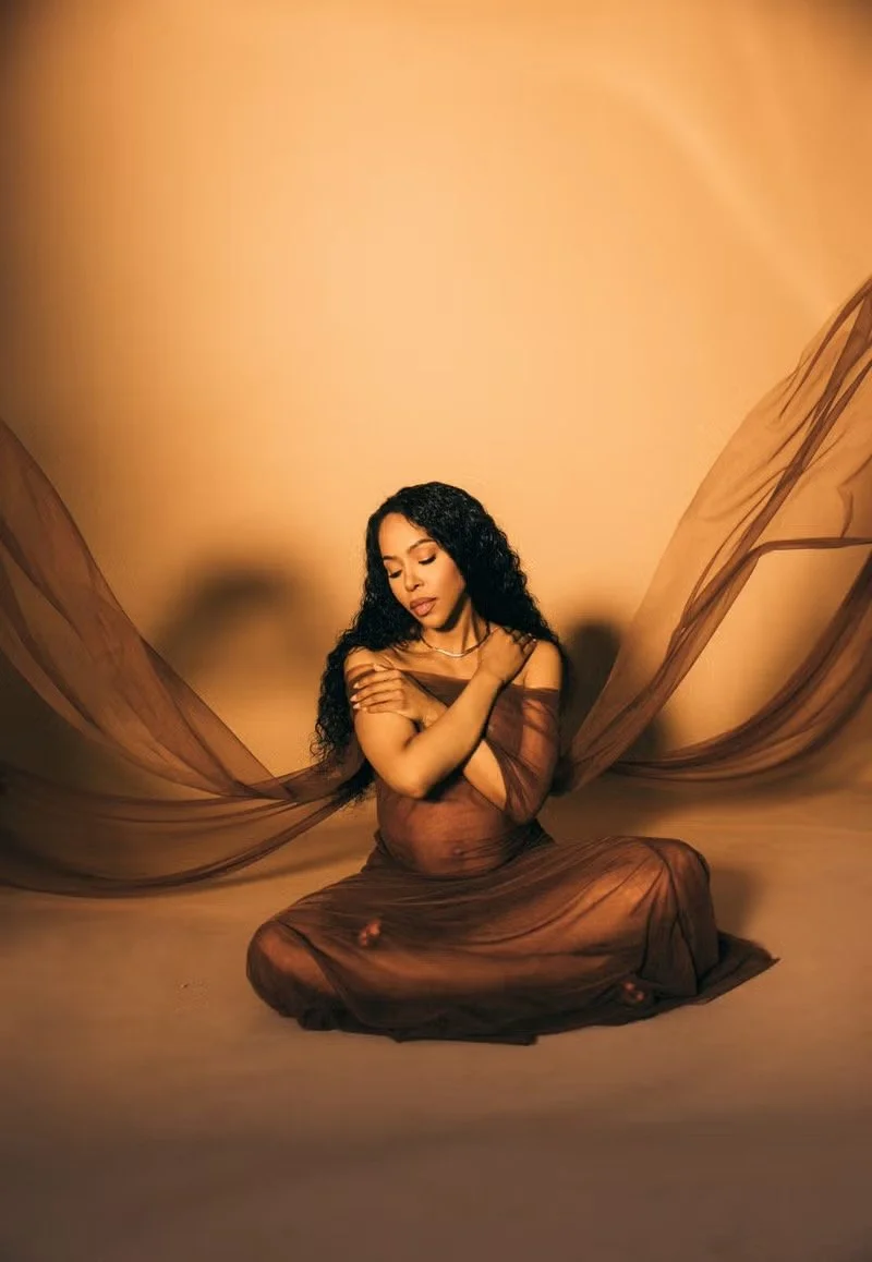 A woman with long curly black hair sitting cross-legged on the floor, dressed in a sheer brown fabric, with matching sheer fabric flowing in the background, and her arms crossed over her chest, eyes closed, against a warm beige backdrop.