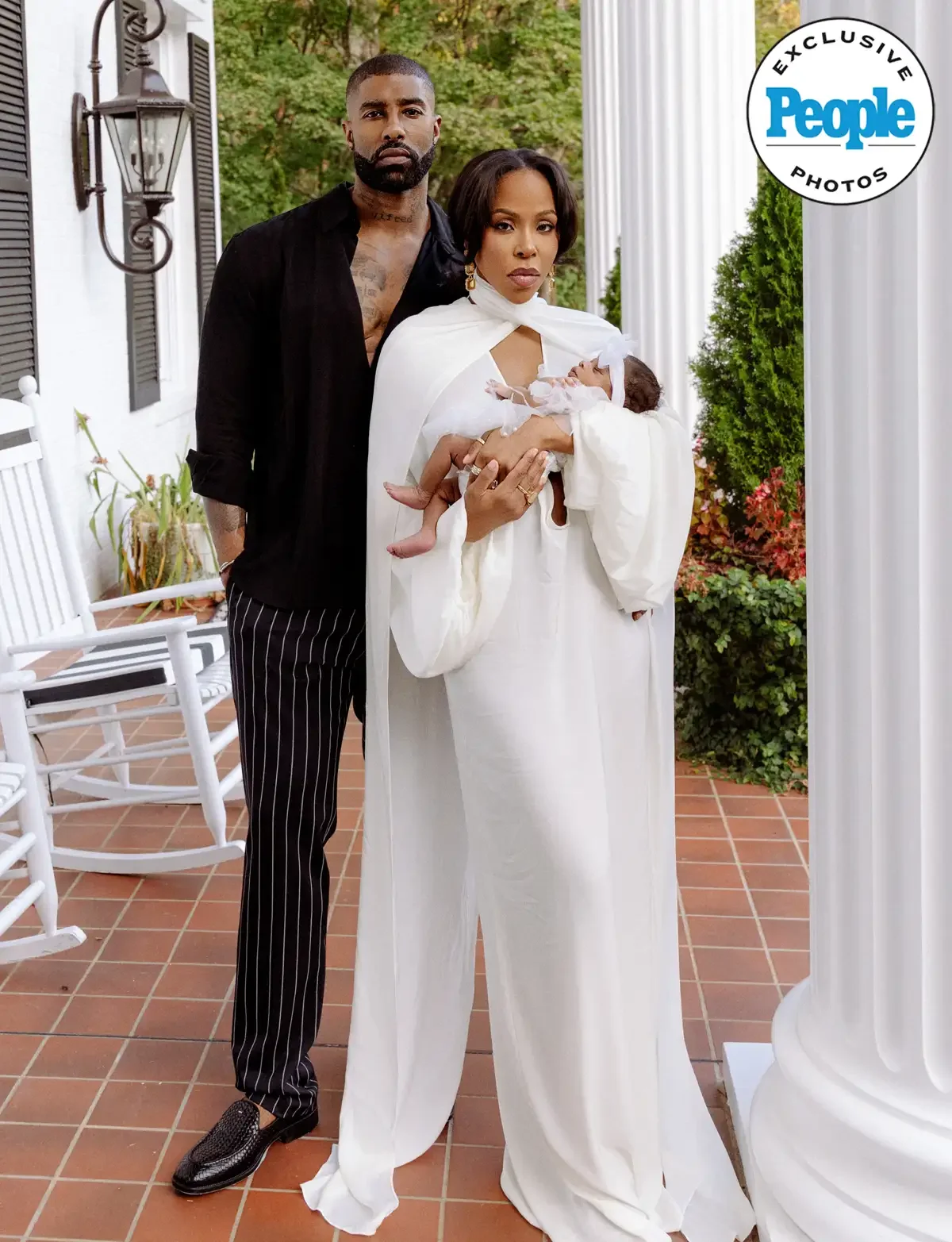 A diverse family of three standing outdoors on a patio with white columns and chairs. The woman, dressed in a white outfit, is holding a newborn baby wrapped in white cloth. The man, dressed in a black shirt and striped pants, stands beside her. They