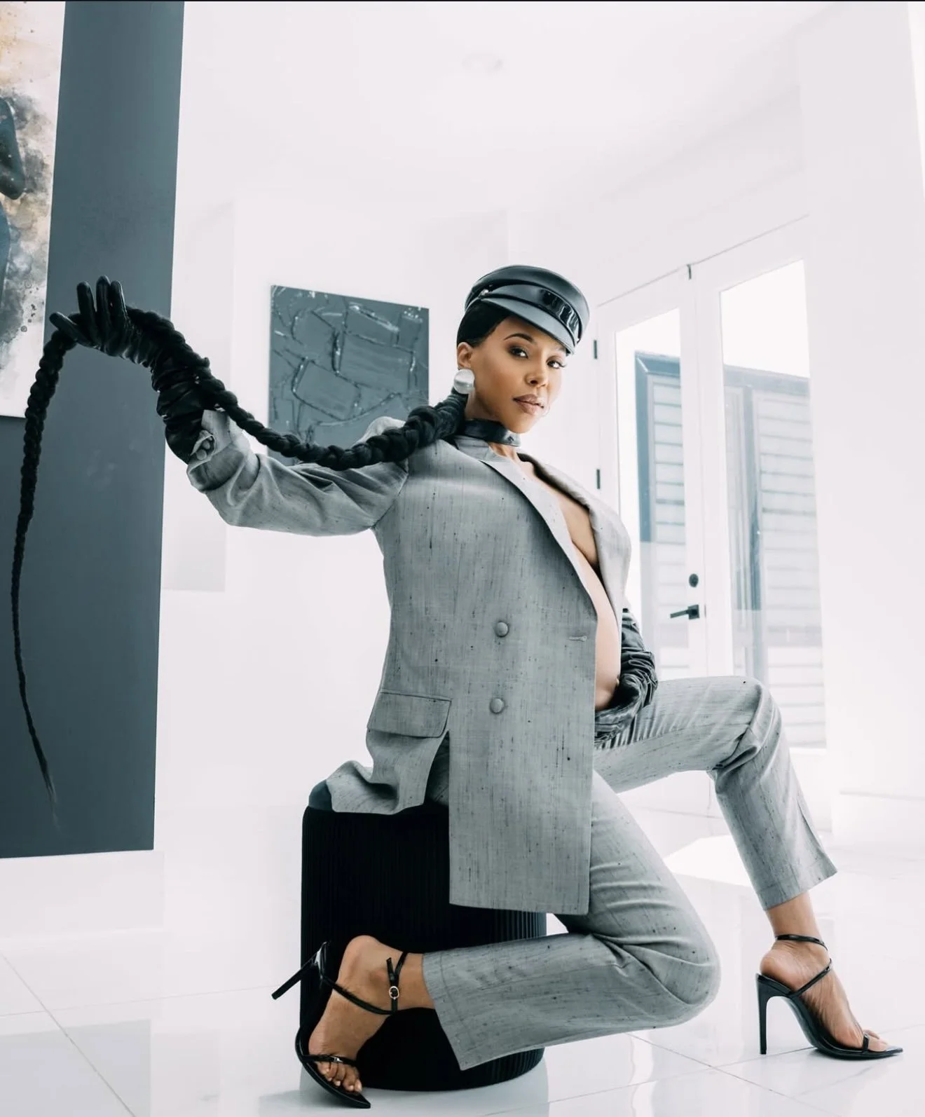 Woman wearing a gray suit and high heels, sitting on a black stool, with one leg crossed over the other, in a bright, modern room with abstract art and large windows.